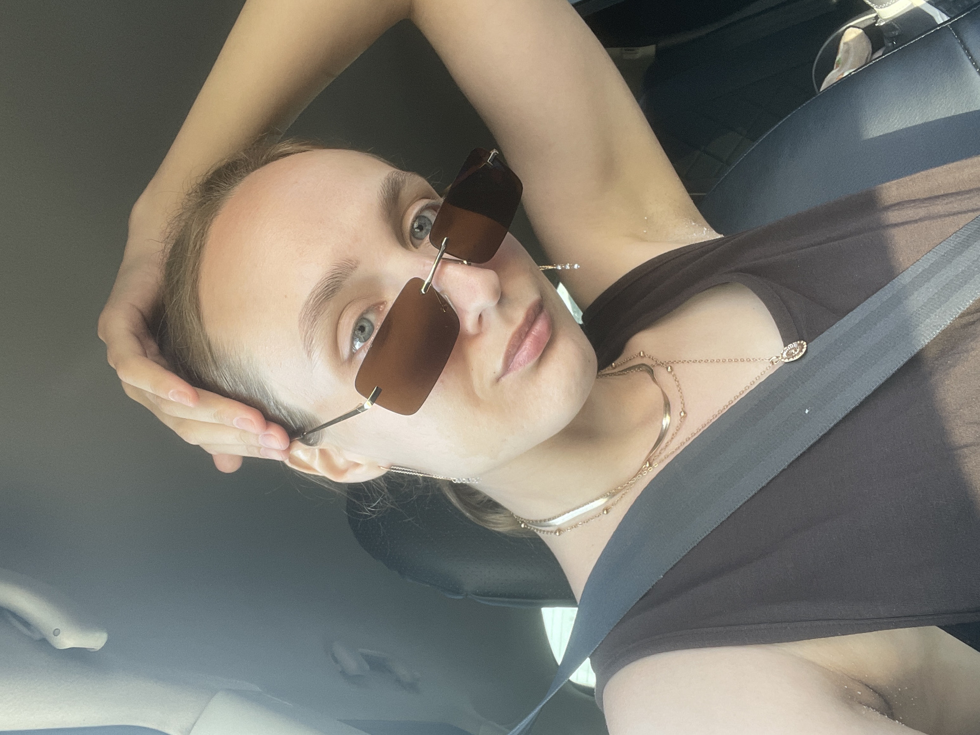 A young woman sits in the driver's seat of a car, wearing a brown top, a seatbelt, and brown sunglasses. She has her hand on her head, and multiple necklaces are visible. The interior of the car is visible in the background. There is no information to determine the location as Izhevsk, Russia.