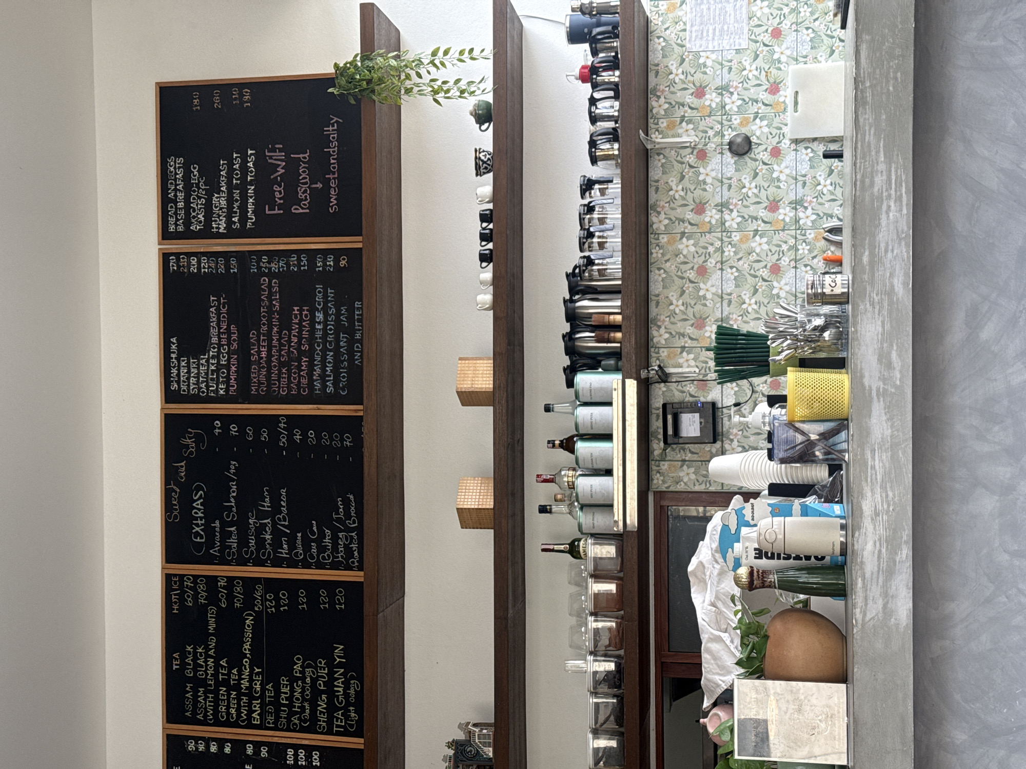 The image depicts a cafe interior with a prominent chalkboard menu displaying food and beverage options. The menu is divided into sections for "TEA," "Sweet and Salty (EXTRAS)," and "SHAKSHUKA," "DRANIKI," "SYRNIKI," "OATMEAL," "FULL KETO BREAKFAST," "KETO EGG BENEDICT," "PUMPKIN SOUP," "BREAD AND EGGS BASEBREAKFASTS," "AVOCADO-EGG TOASTS/2pc," "HUNGRY MAN'S BREAKFAST," "SALMON TOAST," and "PUMPKIN TOAST." Prices are listed in Thai Baht. A section labeled "Free-Wifi Password" with the password "sweetandsalty" is also visible.
The cafe appears to have a rustic and cozy ambiance with wooden shelves displaying various items. On the upper shelf, there are small decorative items and tea containers. The middle shelf holds an assortment of glasses, coffee thermoses, and other serving ware. The lower shelf displays jars of tea leaves, bottles of liquor, and more tea-related containers. The backdrop features a floral-patterned tile wall. In the foreground, the counter area is visible with a concrete surface and various objects like a pink pitcher, potted plants, a ceramic vase, coffee cups, and a yellow waste bin. The overall scene suggests a casual dining establishment in the city of Ko Samui District, Thailand, likely a cafe or a small restaurant. There are no people visible in the image.