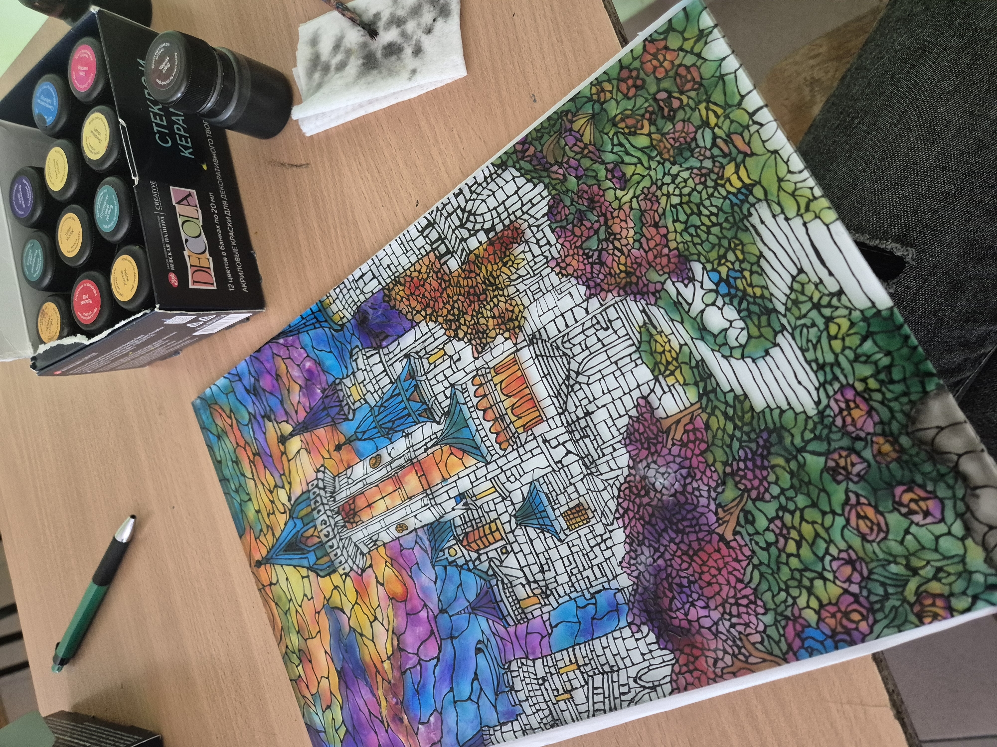 The image presents a crafting scene on a light brown wooden table. A partially opened box of "DECOLA" acrylic paints sits on the left, next to a black jar. Above the box lies a small, soiled white cloth and a green and black pen.
Occupying the majority of the table is a painting of a fantastical castle scene, reminiscent of stained glass. The castle has multiple towers, arches, and a complex network of white lines dividing the picture into small segments, filled with a variety of colors, including blues, purples, yellows, oranges, greens, and reds. The surrounding area features a dense, colorful garden or forest with vibrant flowers.
The image does not appear to be taken in Pavlikeni, Bulgaria.