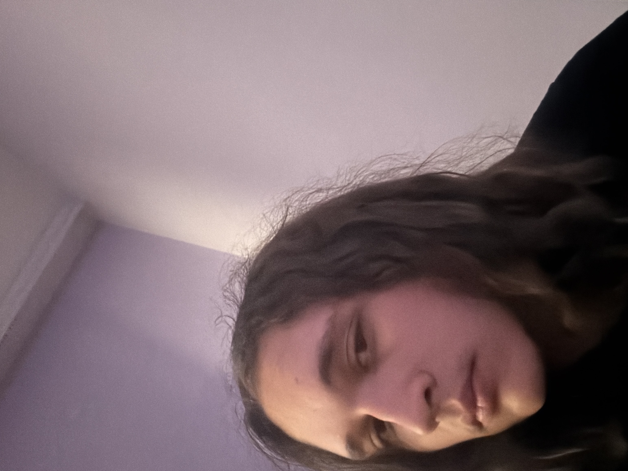 A person with dark hair is angled downward in the frame. The person's face is visible, showing a nose, eye, and lips. The background includes a white and lavender surface, potentially a wall or ceiling. The person is wearing a dark-colored garment. There is no evidence to suggest this image was taken in Asenovgrad, Bulgaria.