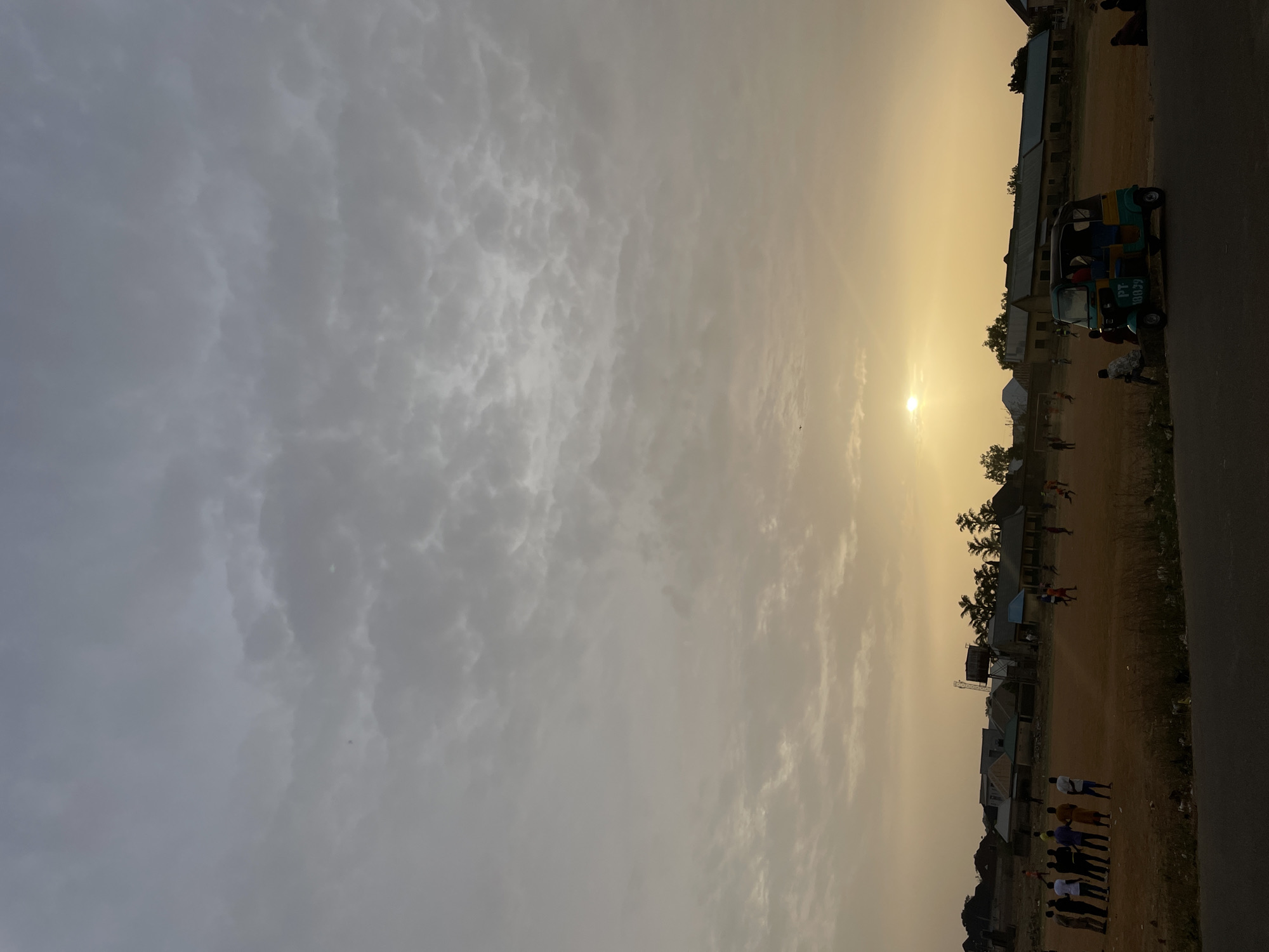 The image shows an outdoor scene in Jos, Nigeria, likely during sunset or sunrise. The sky is partially cloudy, with the sun shining brightly. A vehicle with the inscription "PT. 80639" is parked beside a building. Several people are present, some gathered in a group and others appearing to be playing a game on a field. The environment includes buildings, trees, and a road. The foreground appears to be a dirt or grassy area.