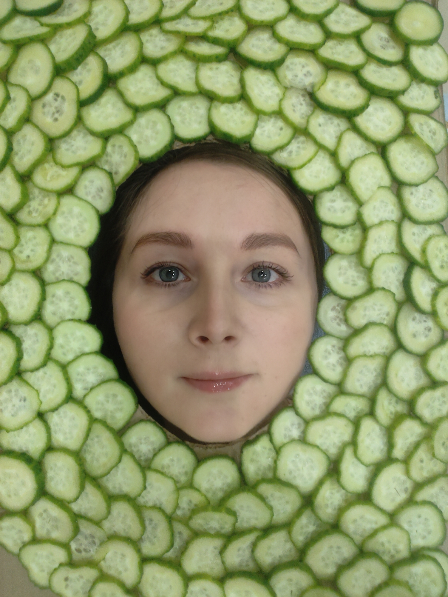 A fair-skinned person with light brown hair is framed by a ring of sliced cucumbers. The person's face is centered in the image, with a neutral expression, blue eyes, and a subtle shine on their lips. The cucumber slices are arranged tightly around their face, creating a green circular border. The background behind the cucumbers is partially visible.
