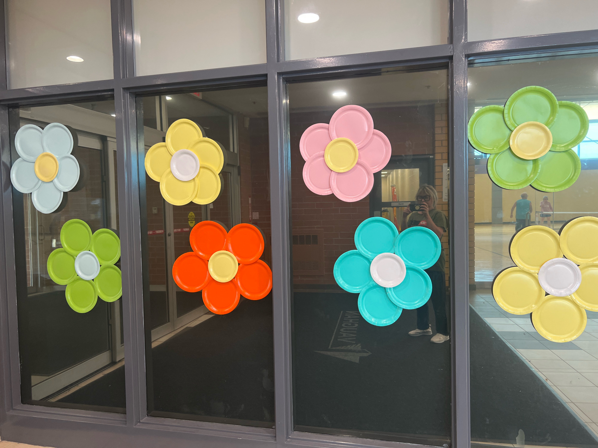 The image shows a storefront window decorated with paper plate flowers. The flowers are arranged in a grid pattern across the glass. There are six flowers visible, each made of seven paper plates arranged in a circular pattern with one plate in the center. The flowers are in various colors including light blue, yellow, pink, light green, orange, and turquoise. A person is visible in the reflection of the window, holding a camera. The storefront appears to be in a commercial area, with a floor mat and doors visible inside. The location is identified as Vaughan, Canada.