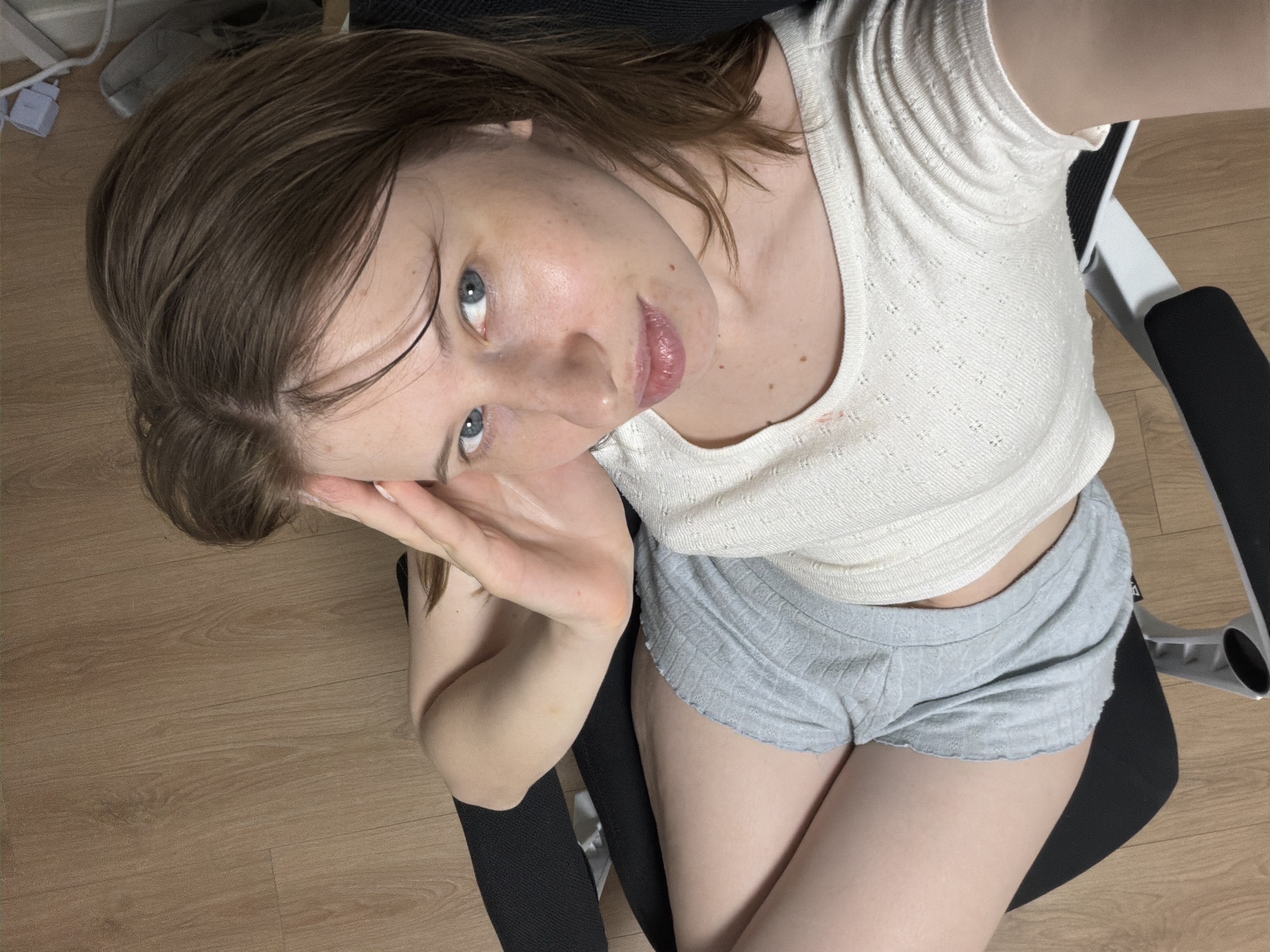 This is a high-angle, medium shot of a young woman seated in a black office chair with white accents. She has fair skin, freckles, and light brown hair that falls around her face. She is wearing a white, textured knit tank top and grey, ruffled-hem shorts. Her right arm is bent, with her hand resting on her forehead. Her gaze is directed upwards and slightly to the right of the camera, with a relaxed and slightly playful expression.
The background consists of a light brown wooden floor and a sliver of a white electrical outlet and cord. The woman is positioned over the seat of the office chair, with the chair's arms and base visible on the right side of the frame. The lighting appears to be from above, creating soft shadows. The overall scene suggests an indoor setting, possibly a bedroom or office. No visible text is present in the image. The time of day and weather are not discernible. The location context places this in Dongducheon-si, South Korea.