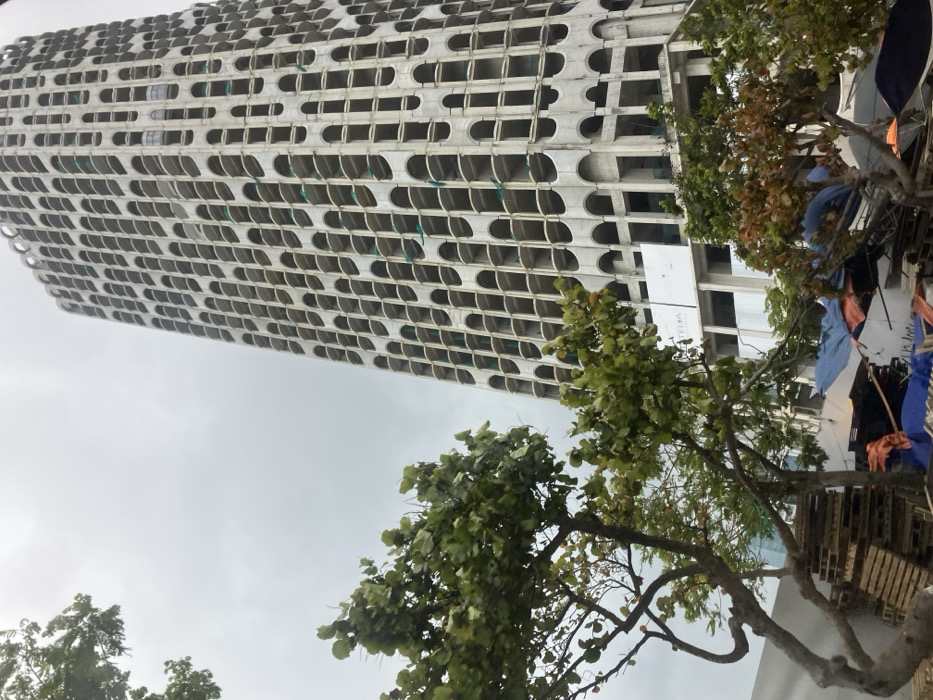 The image presents an urban landscape in Hải Vân Ward, Vietnam, under a uniformly overcast sky. A tall, slender concrete high-rise building, seemingly incomplete or under construction, occupies the majority of the right side of the frame. Its facade is characterized by a repetitive grid of numerous elongated, arch-shaped openings, which serve as window frames, revealing interior structural elements. Sections of green safety netting are visible within some of these openings. A white banner with "TELIA" visible in black text is affixed to a lower section of the building's exterior. The foreground and midground on the left feature multiple trees with green foliage. Below these trees, an area contains stacks of wooden pallets and various items covered by blue and orange tarpaulins, suggesting a makeshift storage or work zone. No individuals, explicit actions, or interactions are discernible within the scene.