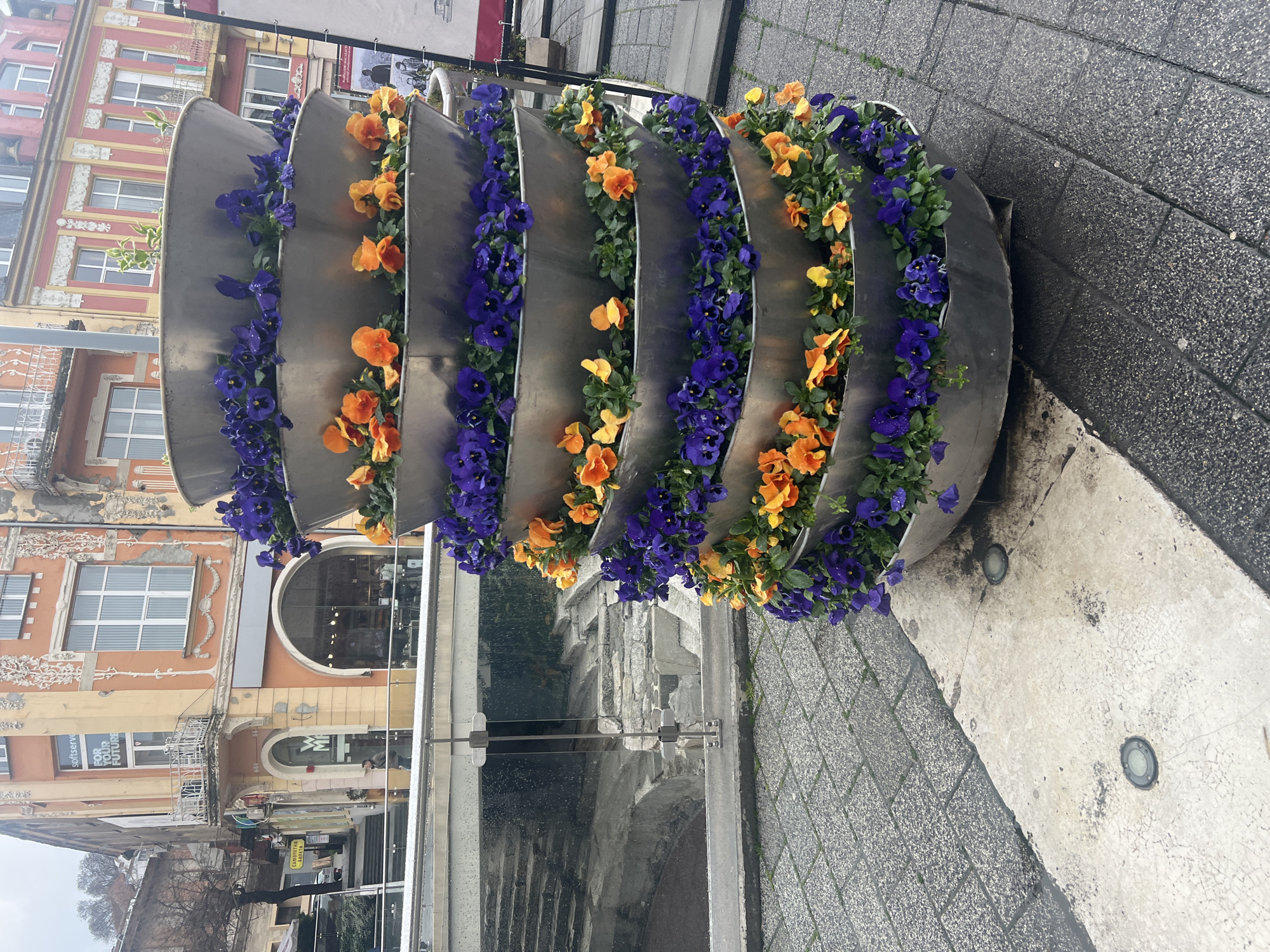 The image shows a spiraling, tiered metal planter filled with orange and purple pansies, positioned on a gray cobblestone sidewalk. In the background, across a glass railing, an archaeological excavation is visible. Behind the planter and the excavation, there are older buildings with decorative facades, typical of urban architecture. A street with shops is visible further down. The location is in Plovdiv, Bulgaria, likely near a public area where historical sites have been uncovered and integrated into the modern city landscape. No people are immediately visible in the frame.
