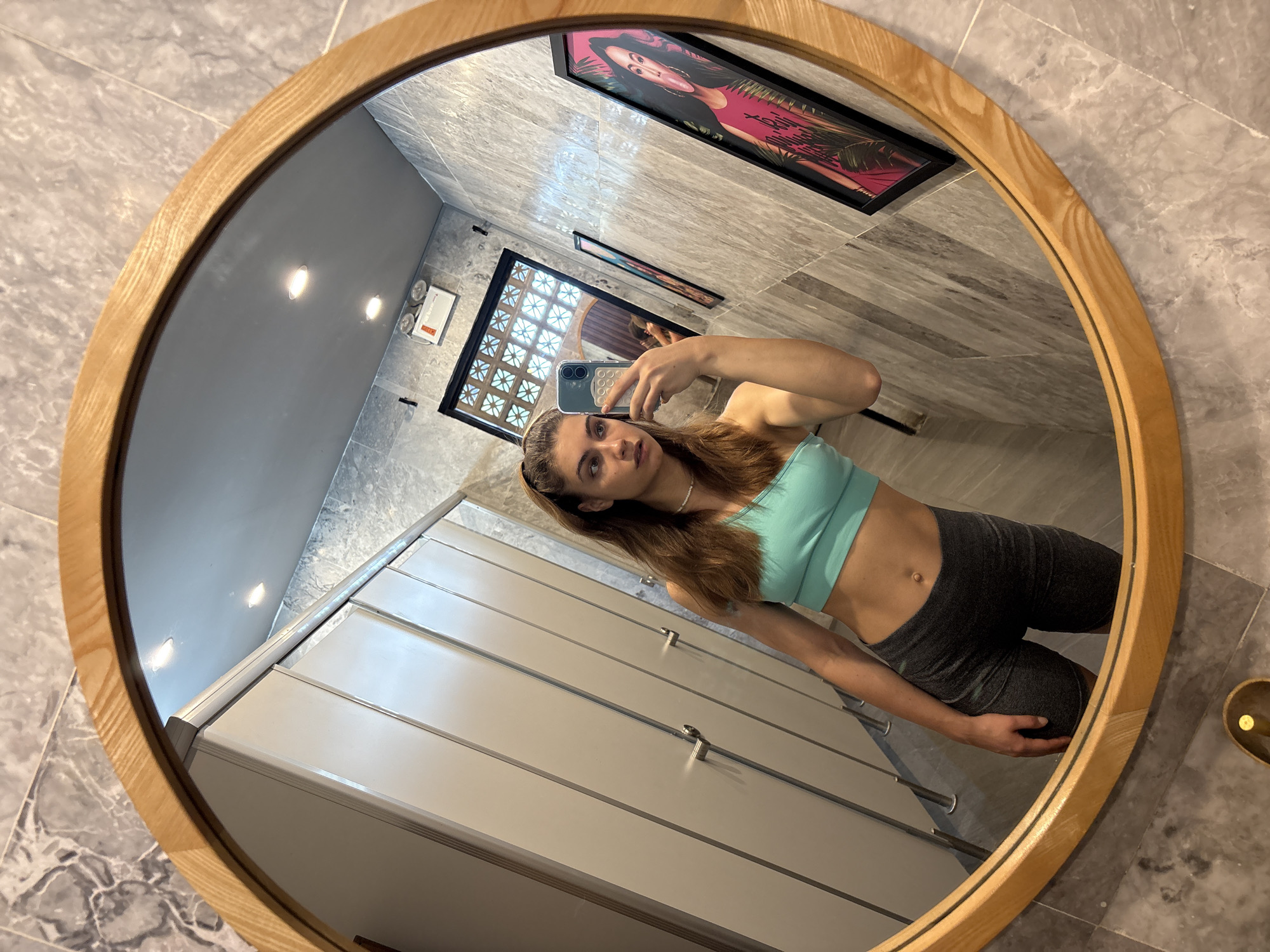 A woman is taking a selfie in a mirror in what appears to be a bathroom. The mirror has a round, wooden frame. The woman is wearing a light green sports bra and dark gray athletic shorts. In the background, there are lockers or storage units with horizontal lines, and the walls are tiled. There are framed pictures on the wall behind the woman. Based on the architectural style visible in the reflection, the location is not likely in the city of Đà Nẵng, Vietnam.