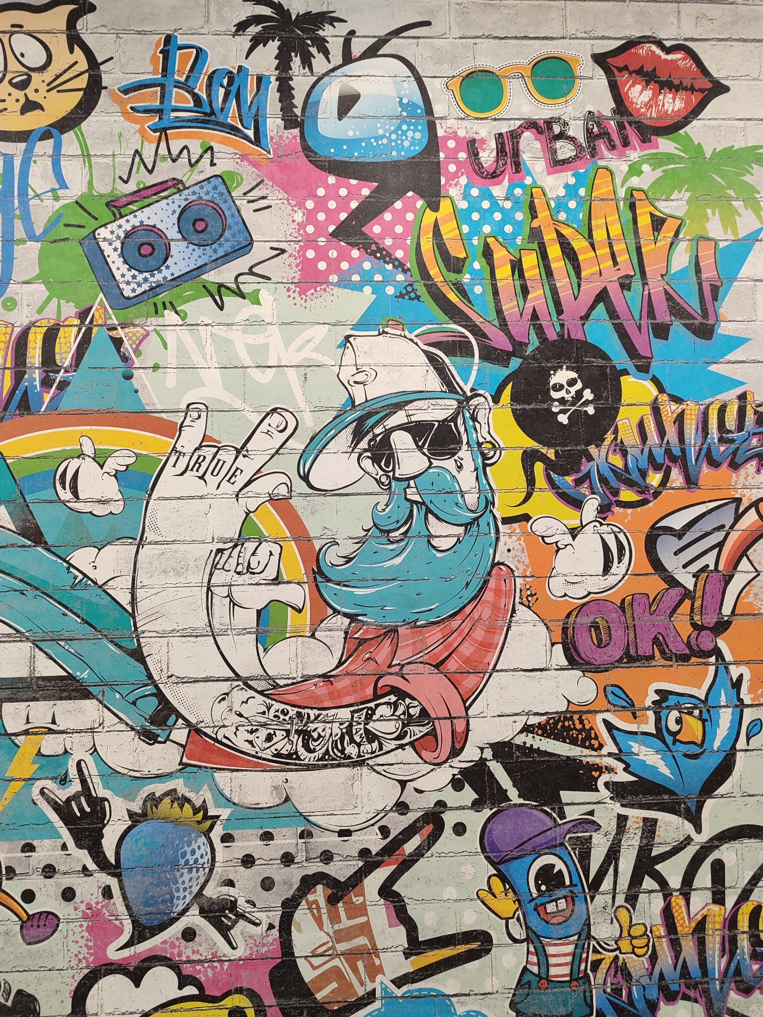 This vibrant and eclectic graffiti mural covers a brick wall, showcasing a chaotic yet energetic mix of cartoon characters, symbols, and text in a distinct pop art and street art style. The mural, located in Izhevsk, Russia, features a kaleidoscope of bright colors including blues, pinks, yellows, greens, oranges, reds, and black and white accents.
The wall is densely packed with subjects. In the upper left, a wide-eyed cartoon cat is depicted near stylized "Boy" lettering, a black palm tree, and an abstract blue character with sunglasses. Below them, a retro boombox with star patterns is surrounded by dynamic lines. The upper right section displays yellow sunglasses, bright red lips, and the prominent word "URBAN" alongside a large, multicolored "SUNDAY" tag. A black circle with a white skull and crossbones is also present.
The central focus is a large, white hand making a gesture, with the word "TRUE" emblazoned across its fingers. Behind it, a rainbow arc adds another layer of color. Adjacent to the hand is a bearded character wearing sunglasses, a cap, and a red scarf or cloak, exuding a cool, laid-back demeanor. Clouds punctuate various elements throughout the composition.
Towards the bottom left, a blue strawberry character with a yellow crown makes a "rock on" hand gesture. Further right, the word "OK!" is written in bold purple letters, next to a blue, bird-like creature. A worm-like character with a purple cap and striped overalls, giving a thumbs-up, anchors the bottom right.
The entire mural is characterized by dynamic shapes, drips, splatters, polka dots, and zigzags, all contributing to an urban and youthful aesthetic. The artwork evokes a sense of rebellious playfulness and modern street culture, without depicting a single specific event or time of day, rather a collection of iconic imagery creating a rich visual narrative.