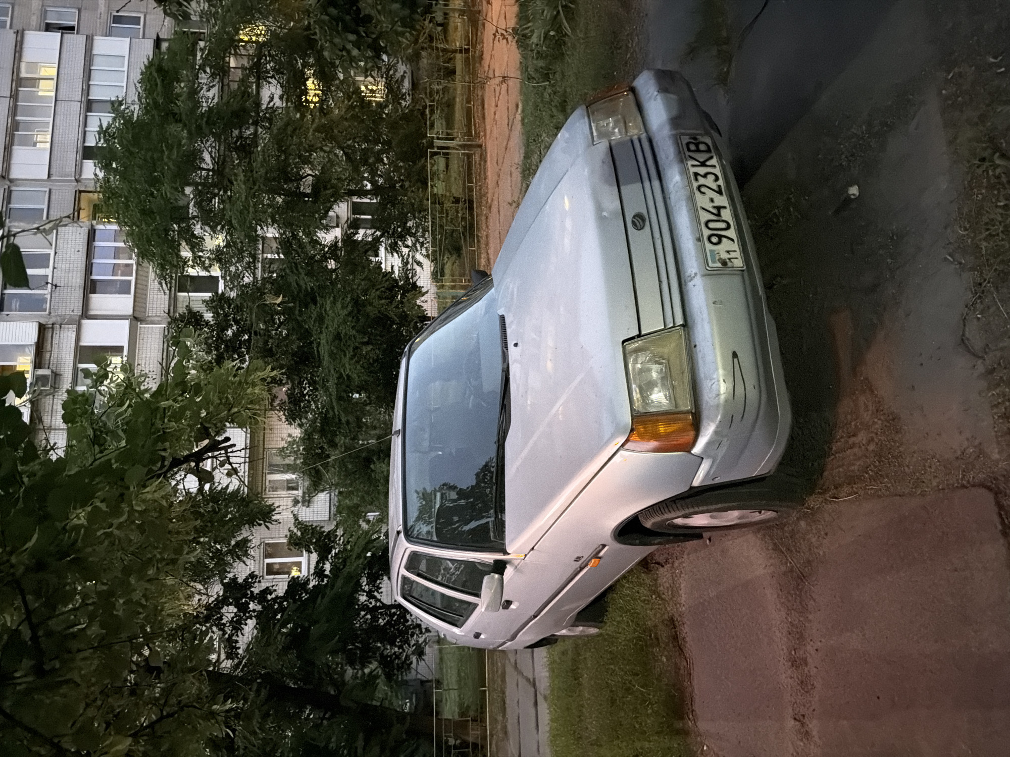 A silver car with license plate "904-23KB" is parked on a paved surface next to a grassy area. A large, leafy tree is on the left side of the car, with a multi-story building in the background. The building has many windows, and some of them are lit, suggesting it is evening or nighttime. The sky is dark, indicating it is likely nighttime. The scene appears to be in a residential area, possibly a courtyard or street. The car is the primary object in focus. There are no people visible in the image. The lighting suggests it is late evening or early night.