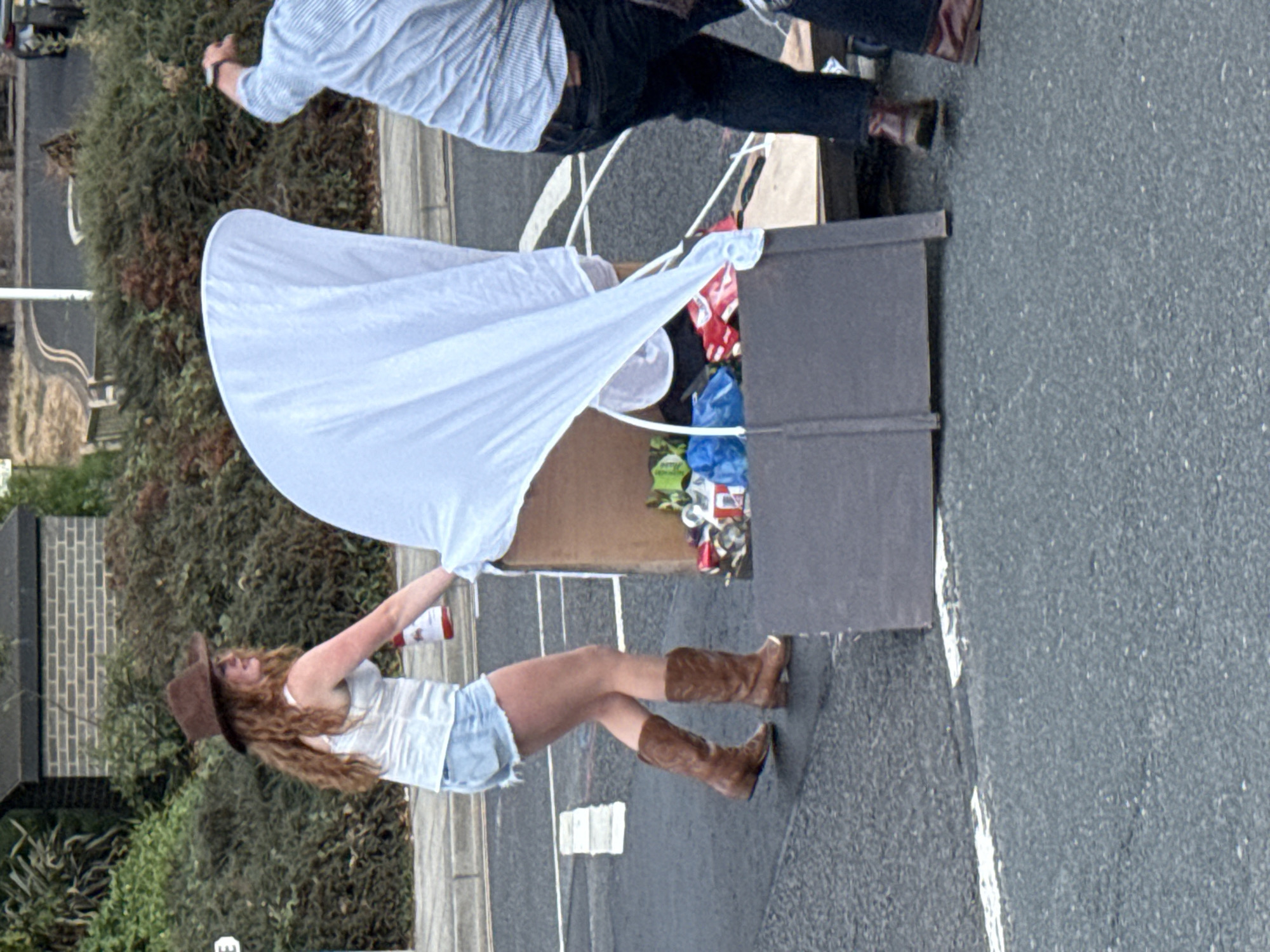 This image depicts a street scene in St Leonards, United Kingdom. A woman wearing a brown cowboy hat, a white tank top, and denim shorts is standing on asphalt. She is holding a red and white object, possibly a bottle, in her right hand and appears to be interacting with a large, brown cardboard structure draped with a white sheet. This structure contains various items, including what appear to be bags and cans, suggesting it might be a makeshift stall or display. Next to the woman, another person, partially visible, is wearing a blue striped shirt and dark trousers, and is also near the cardboard structure. The background shows greenery, a brick building, and a street with white markings.