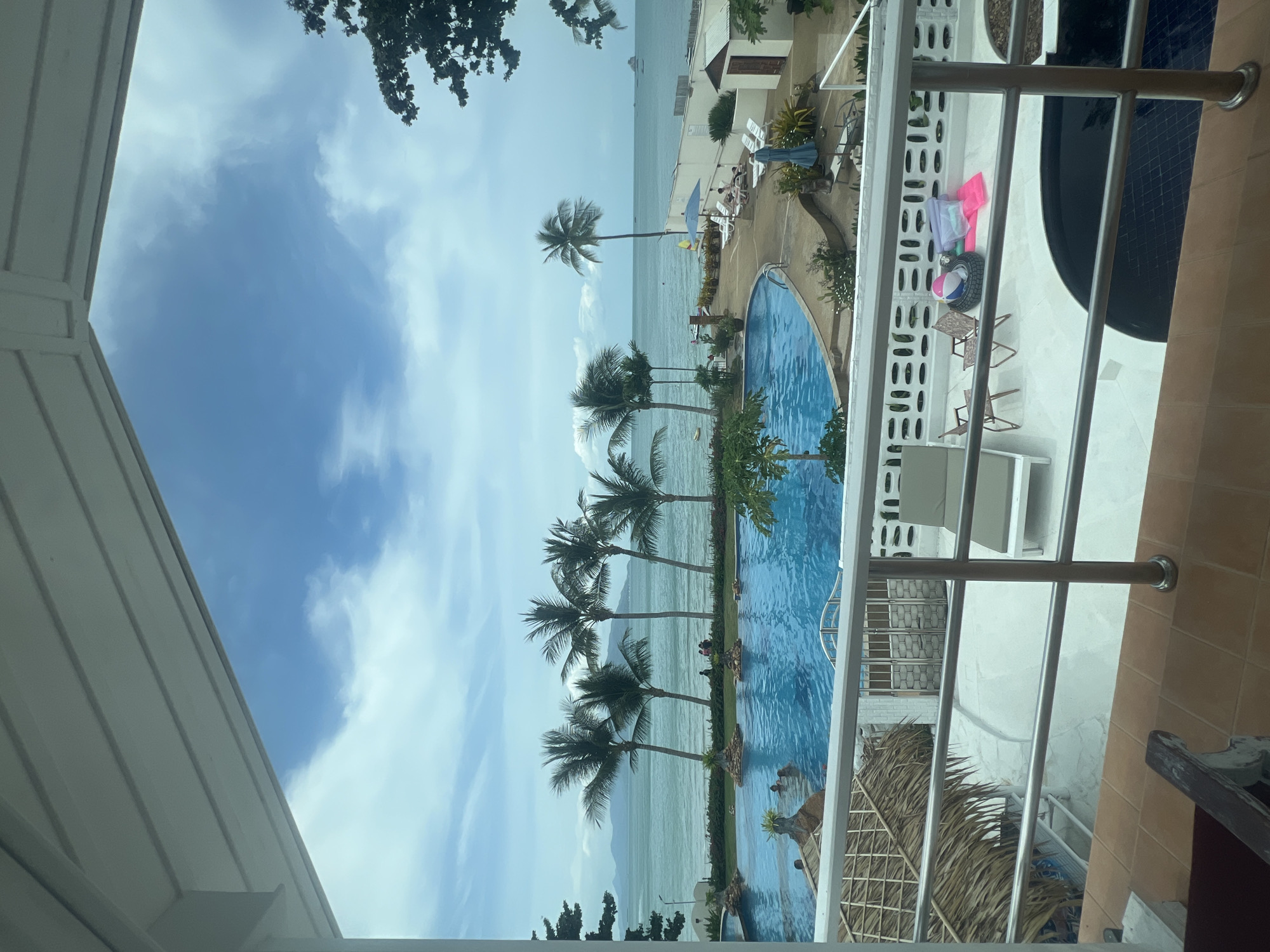 A view from a balcony overlooks a resort with a swimming pool, the sea, and a cloudy sky. In the foreground, a white balcony railing separates the viewer from the resort. Beyond the railing, a large blue swimming pool is filled with people, and several palm trees line the edge of the pool and extend towards the ocean. The sea stretches out to the horizon, with islands visible in the distance. The sky is partly cloudy, with patches of blue showing through. The resort appears to be located in a tropical coastal area.