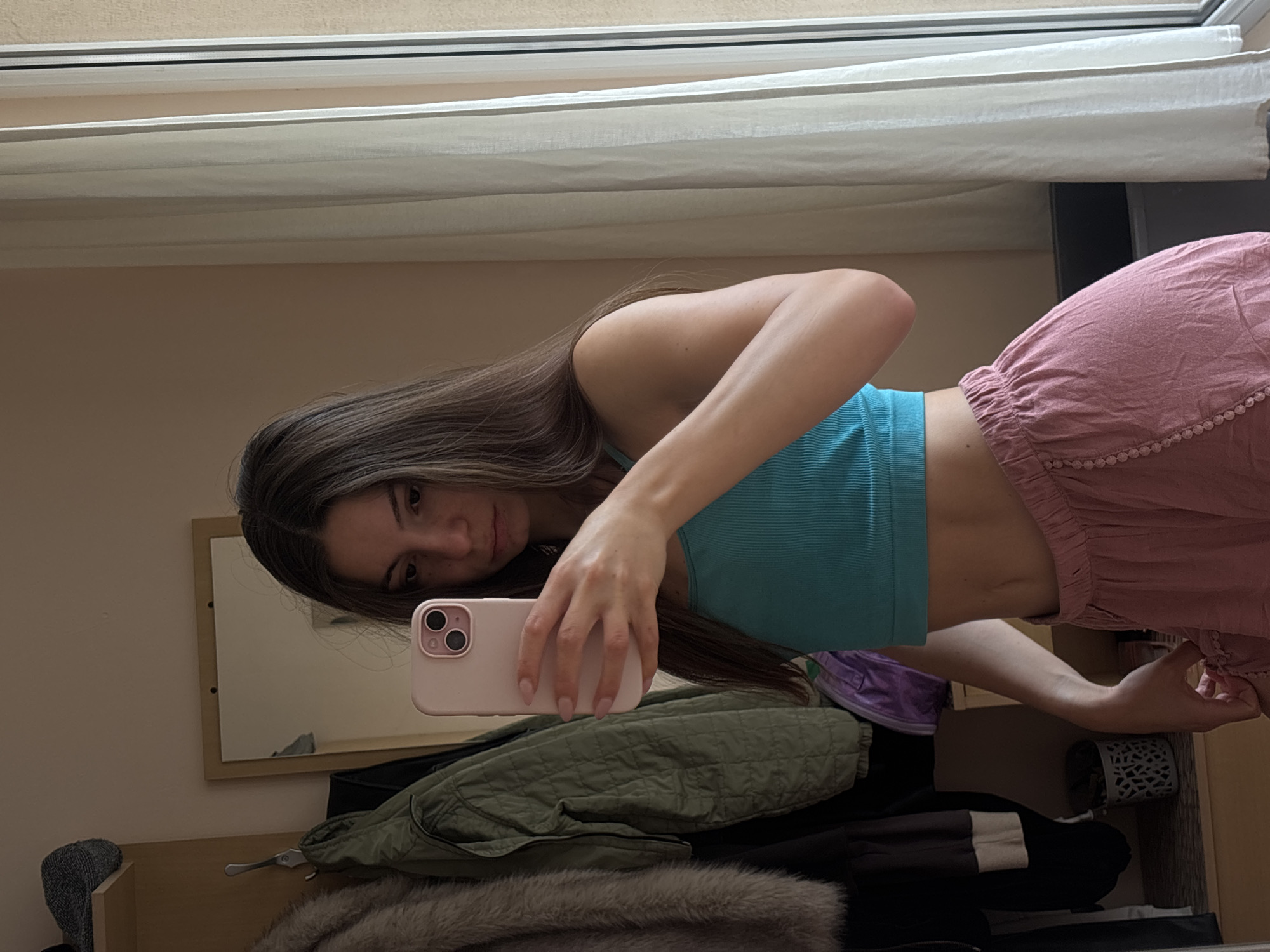 A young woman with long brown hair is taking a selfie in a mirror. She is wearing a turquoise crop top and pink pajama-like shorts. She is standing in what appears to be a bedroom, with a closet visible to her left and a window with white curtains to her right. The room has neutral-colored walls. The woman is holding a pink phone in her right hand, using it to capture her reflection. The scene is indoors and appears to be daytime, with natural light coming from the window. The woman's expression is neutral.