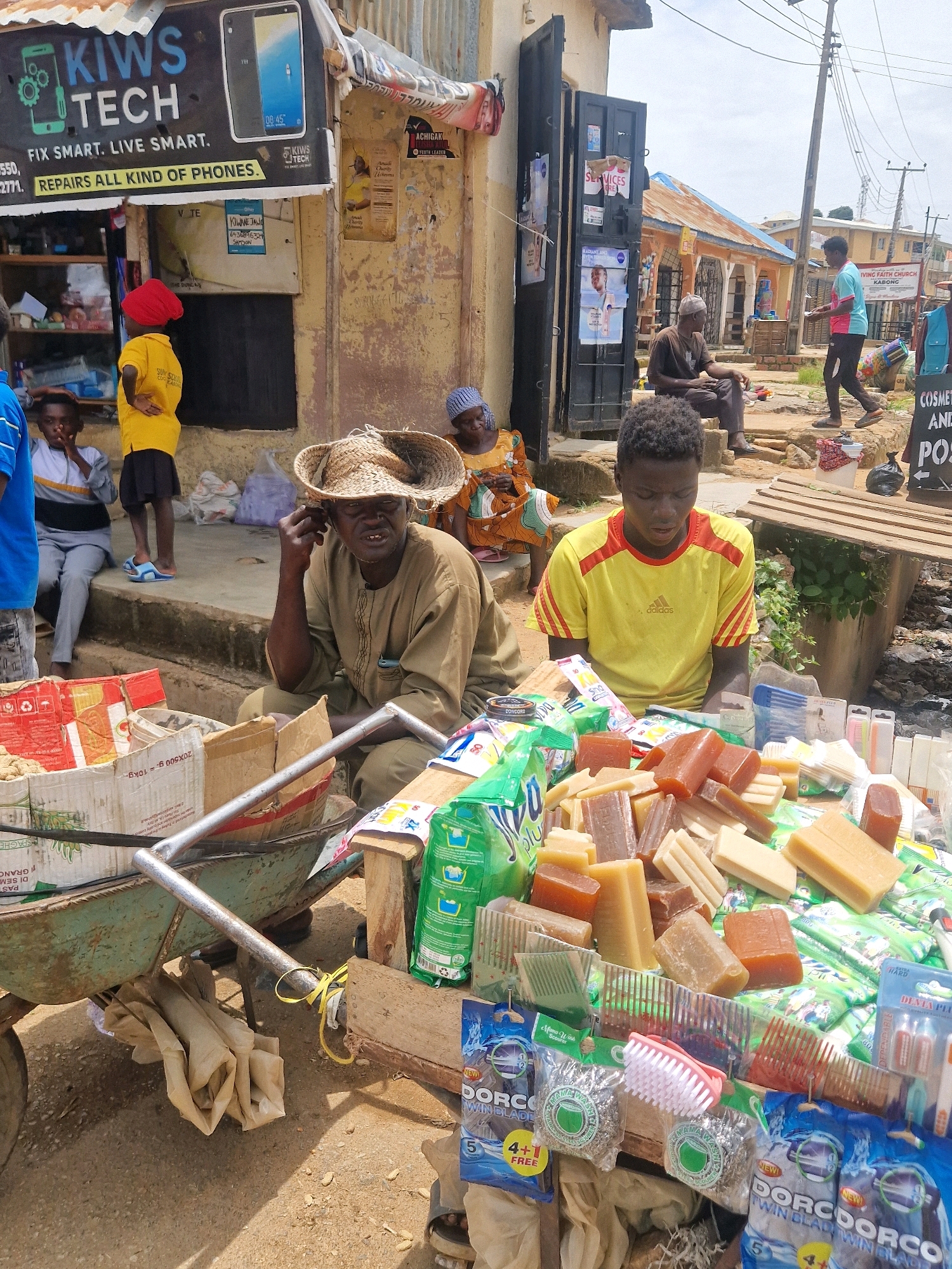In an outdoor marketplace setting, a group of individuals are gathered near a storefront displaying a sign for "KIWS TECH." In the foreground, a vendor sits behind a display of bar soaps and toiletries, including combs and small brushes. To the left, a green wheelbarrow is partially visible, laden with boxes. In the middle ground, another person wearing a straw hat is seated. Behind them, several other individuals, including a child in a yellow shirt and red headscarf, are present. Further in the background, more people are visible near other stalls and buildings, suggesting a bustling commercial area. The ground is paved with dirt and concrete.