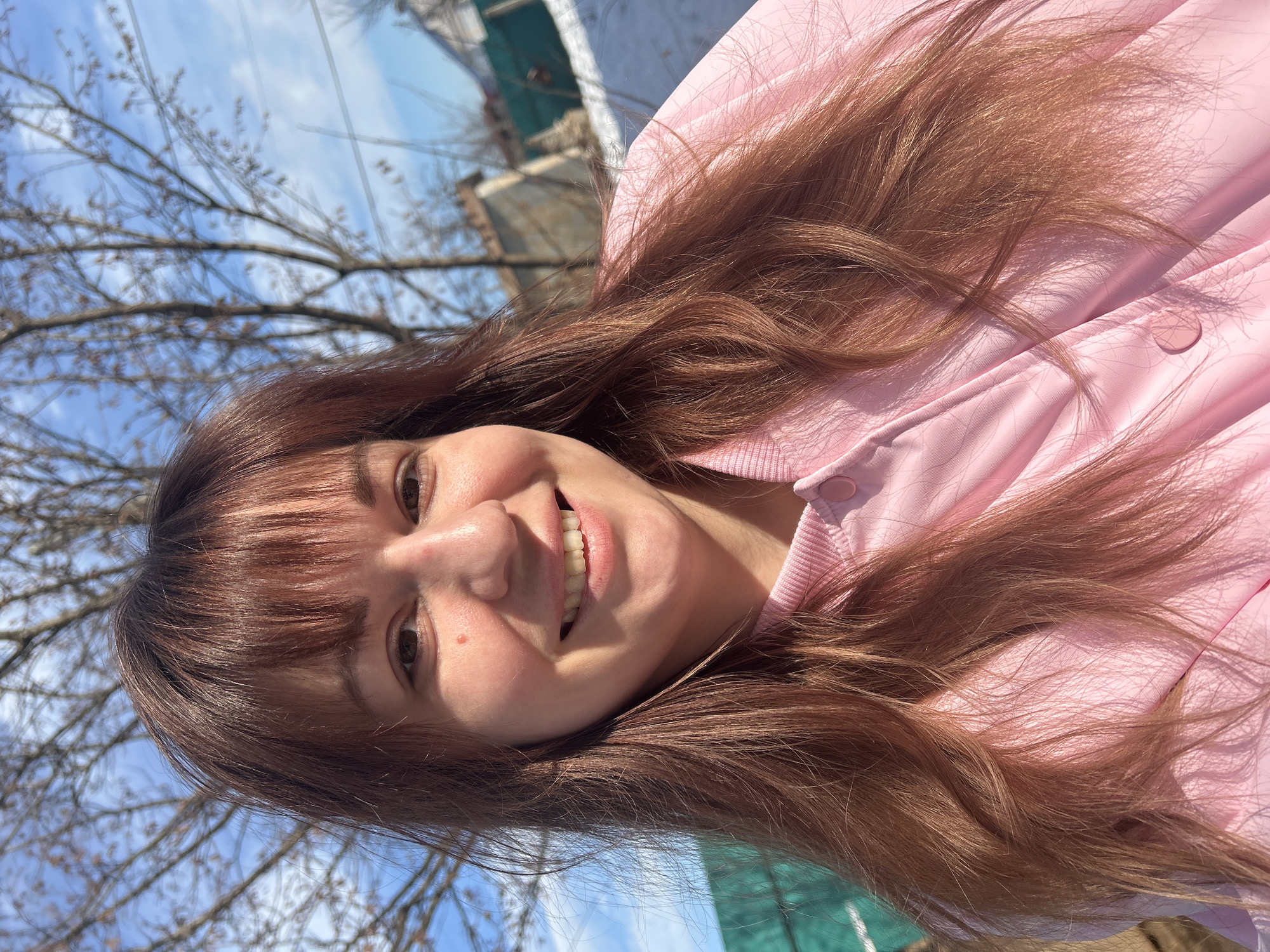 The image displays a head-and-shoulders portrait of an adult female in an outdoor setting in Фоки, Russia. The subject has long, brown-reddish hair with bangs across the forehead and is wearing a light pink collared jacket. She is smiling, revealing teeth, and looking directly into the camera. A small reddish mark is visible on her right cheek. The background consists of a clear blue sky with faint clouds and multiple bare deciduous trees. To the left, a section of a green mesh fence is partially visible. To the right, a light-colored building with a visible roof, another green fence structure, and a patch of snow are present. The scene is illuminated by strong natural sunlight.