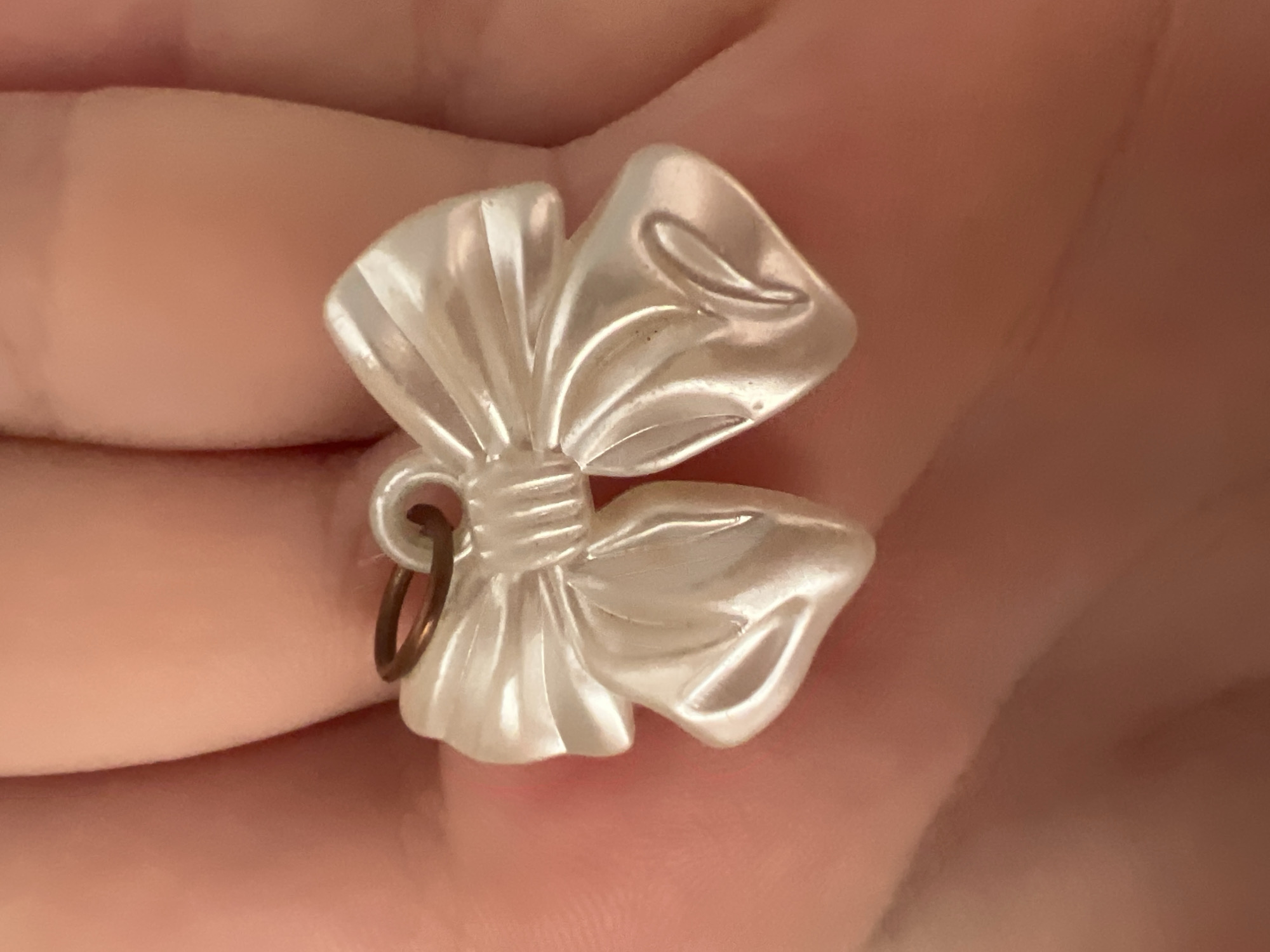 A close-up, eye-level shot captures a pearlescent white bow-shaped charm, likely made of plastic, held between the fingers of a person. The charm is attached to a small, copper-colored jump ring. The fingers are light-skinned and are positioned in the foreground, with the bow slightly out of focus against the softly blurred background of more fingers. The lighting is warm and diffuse, casting subtle shadows on the charm and skin. No specific city or location information is discernible.