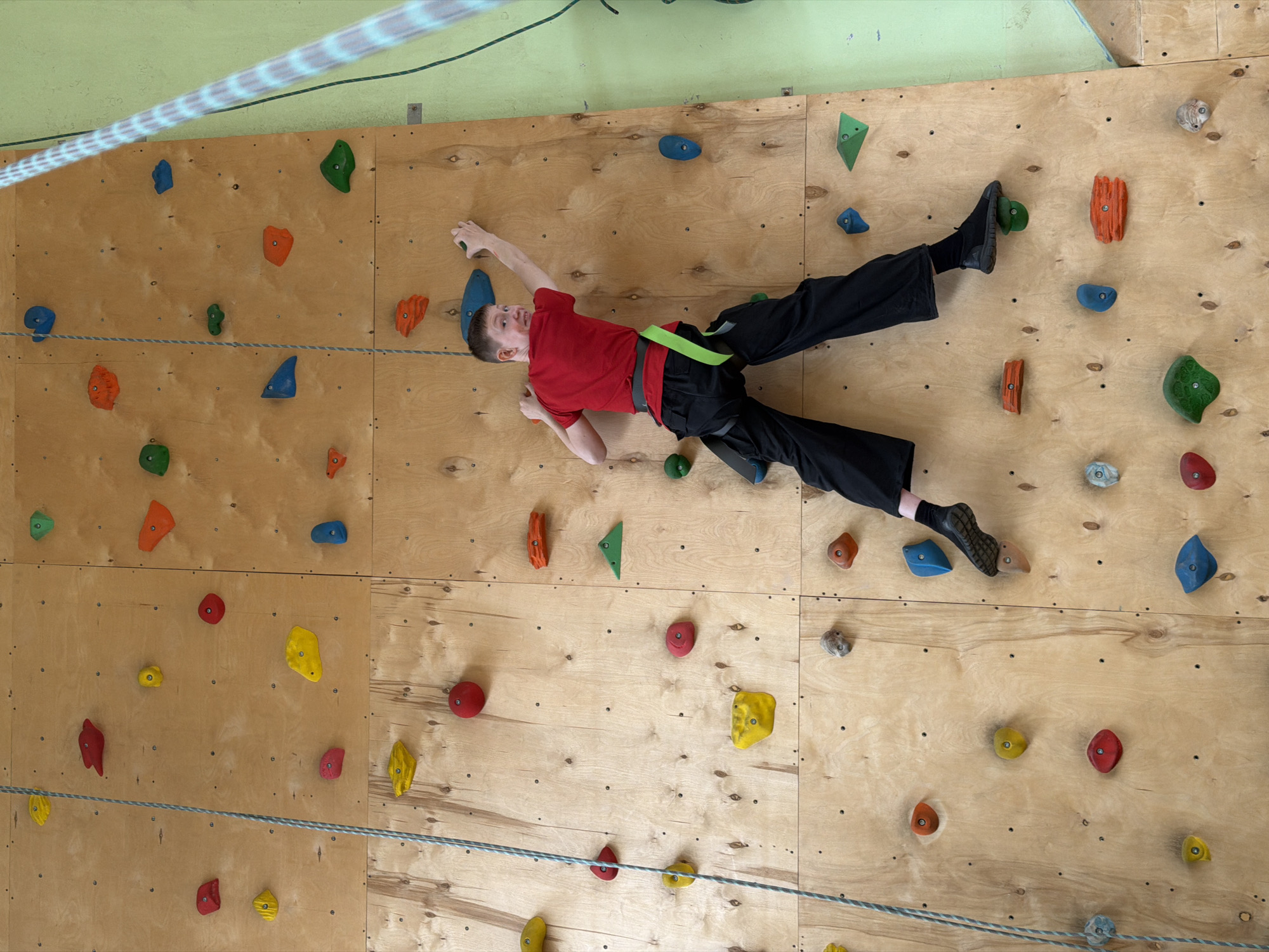 This image, taken in Izhevsk, Russia, depicts an indoor climbing facility. The primary subject is a young male, likely a boy or teenager, actively climbing a wooden bouldering wall. He is wearing a red short-sleeved shirt, black long pants, and a bright green climbing harness. His body is oriented horizontally across the wall, demonstrating a traverse or bouldering movement, with his hands gripping and reaching for various climbing holds and his feet pressed against others. His facial expression conveys concentration and effort.
The scene is dominated by the climbing wall, which is constructed from multiple light-colored plywood panels. These panels are dotted with numerous small screw holes and feature a wide array of brightly colored climbing holds in various shapes and sizes, including blue, green, orange, red, yellow, and grey. Above the wooden wall, a section of a light green wall or ceiling is visible.
Two ropes are present in the image. One, appearing in the upper left, is a thick, light-colored rope with a distinct striped pattern, stretched across the top corner of the frame. The second rope, a thinner grey and white braided line, runs horizontally across the lower half of the climbing wall, possibly serving as a guide, boundary, or part of a safety system. The lighting is bright and even, typical of an indoor facility. There is no visible text in the image.