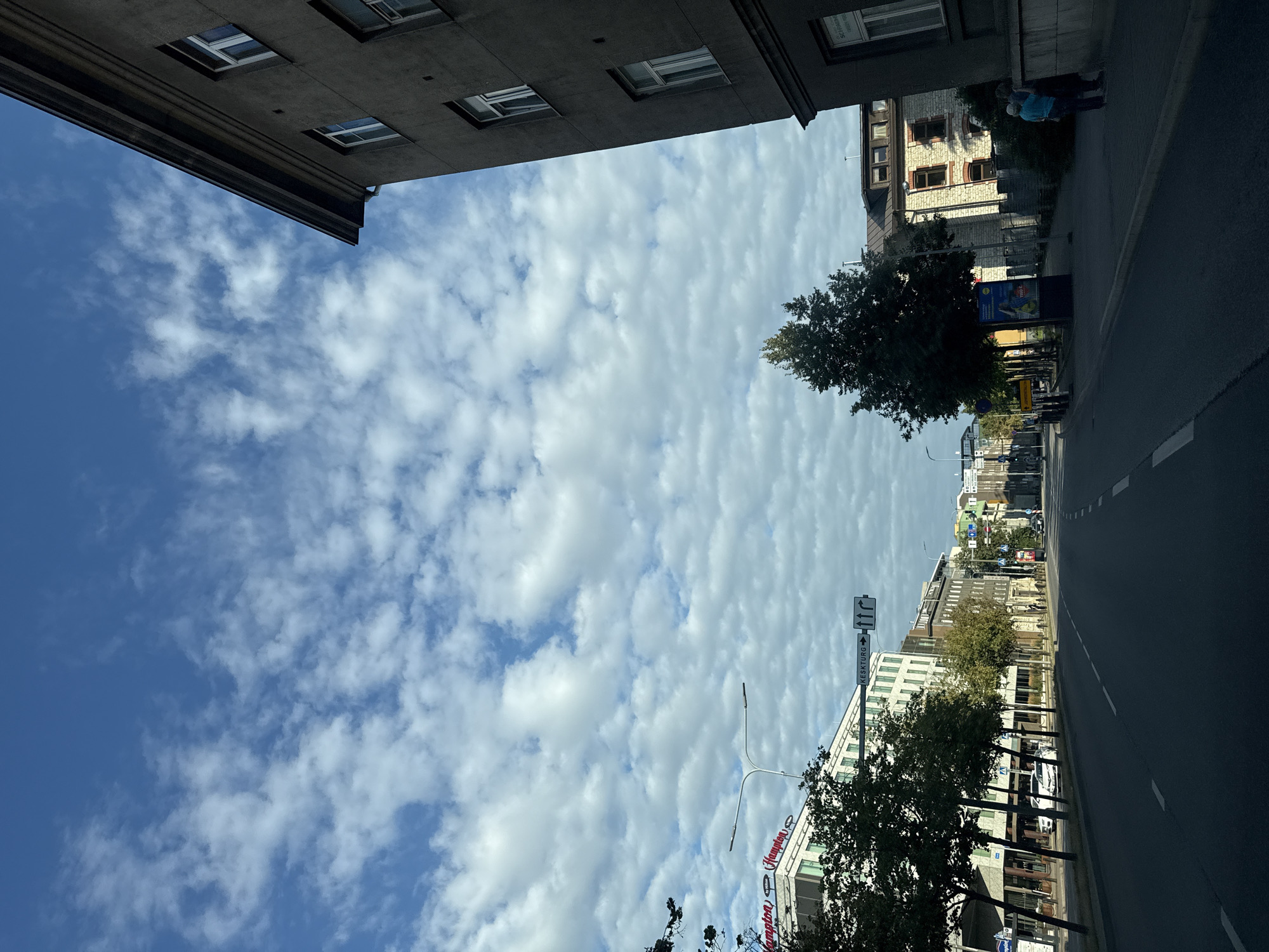 The image shows a street scene in Tallinn, Estonia, with a road in the foreground and buildings on either side. The sky is filled with scattered white clouds against a bright blue background. On the left side of the road, there is a building with the "Hampton" logo visible, and trees line the sidewalk. Further down the road, there are more buildings of various architectural styles, some appearing older and some more modern. Traffic signs are visible, including one indicating "KESKTURG" with arrows. On the right side of the road, a large, dark-colored building dominates the view, with several windows visible. A few people are seen on the sidewalk near this building. The road appears to be a multi-lane urban street with some cars parked and others driving. The overall impression is that of a sunny day in a European city.