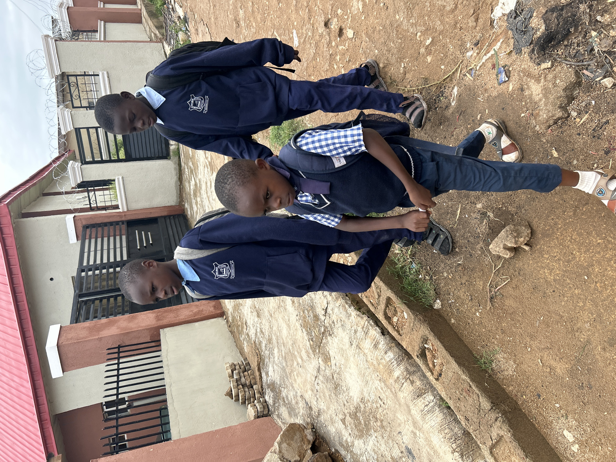 Two boys in school uniforms walk on a dirt path. The path is bordered by a concrete curb and a building with a red roof and tan walls. Debris litters the path. The location is possibly in Jos, Nigeria.