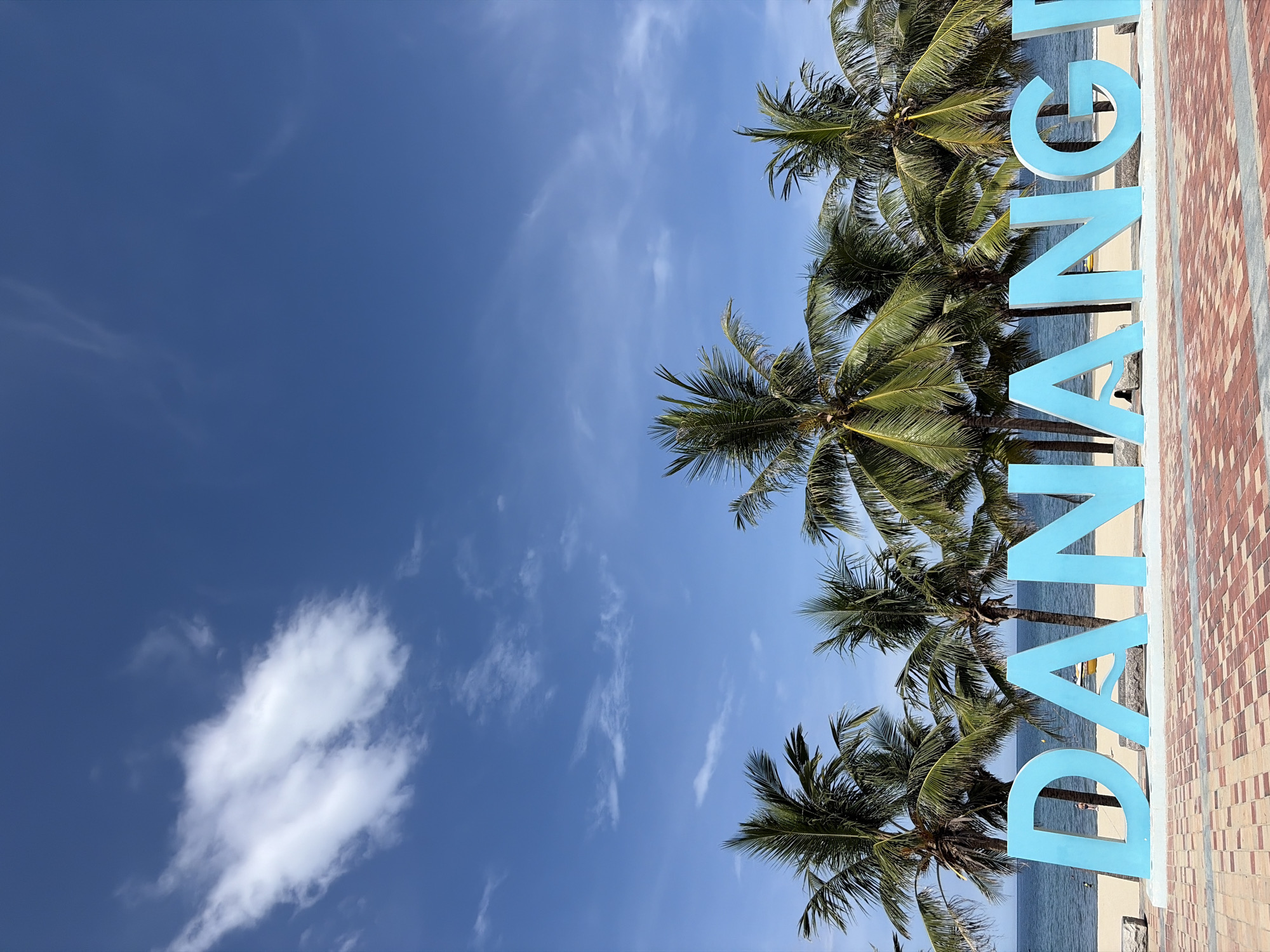 This is a vertical, eye-level, medium shot of a beach sign in Da Nang, Vietnam, on a clear day. In the foreground, large, bright blue letters spell out "DANANG" in all caps. The letters are bold, three-dimensional, and stand on a white base. The ground in front of the sign is paved with red and beige interlocking bricks. Behind the sign, a line of palm trees with lush green fronds stands tall against the ocean. The ocean is a deep blue, with small waves rolling onto a sandy beach. A few scattered clouds, some wispy and thin, others more substantial, dot the bright blue sky. A person is visible on the beach to the left, and a yellow object, possibly a small boat or kayak, is situated on the sand near the water's edge.