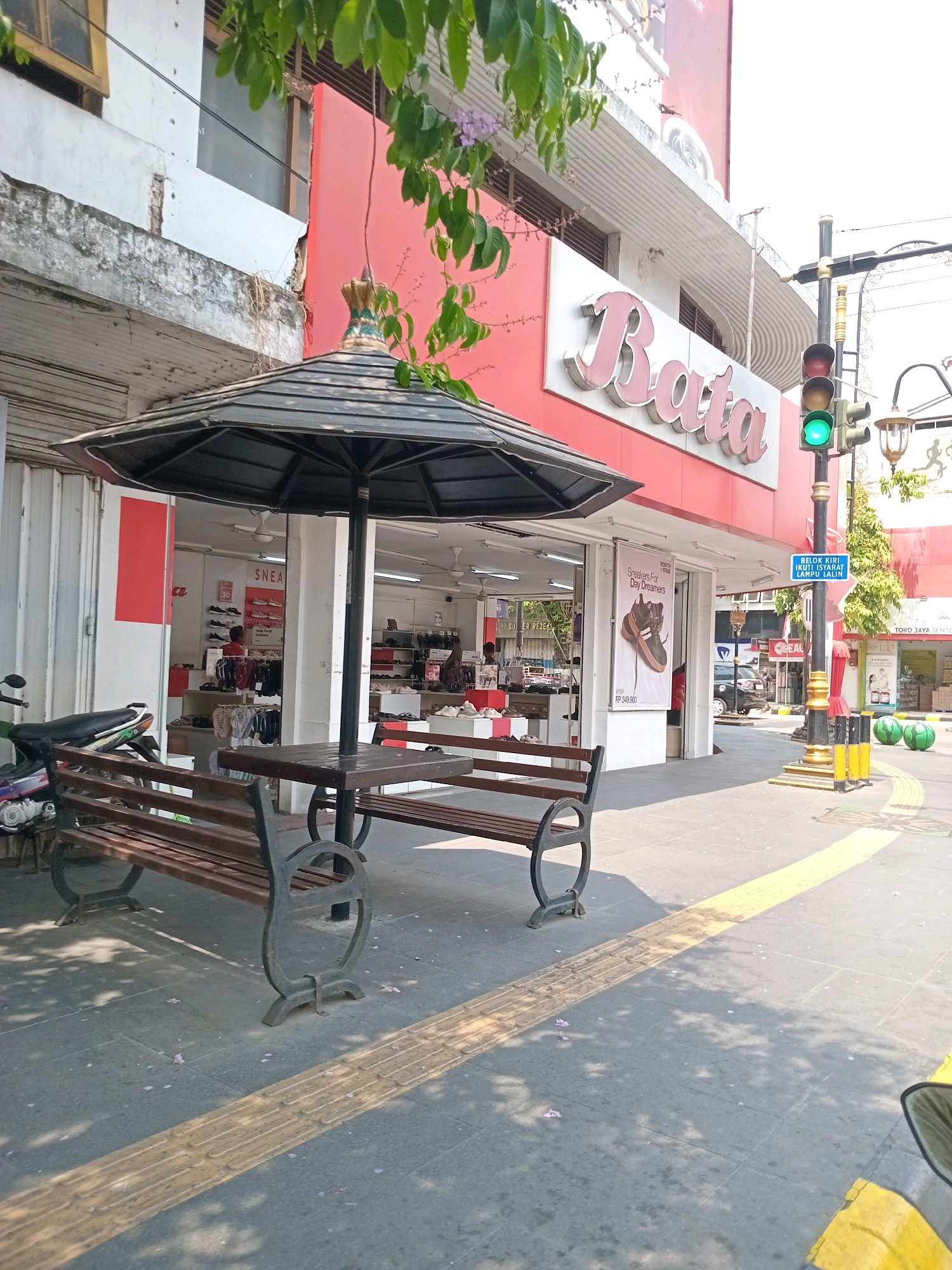 The image depicts a street scene in front of a Bata shoe store. In the foreground, there are two wooden benches and a wooden table under a large black umbrella. A motorcycle is parked to the left of the benches. The Bata store is visible behind the furniture, with its signage prominently displayed. Inside the store, shelves are stocked with shoes, and a few people can be seen. A traffic light with a green signal is on the right side of the image, along with a blue street sign. The sidewalk is paved with grey tiles and has a yellow tactile paving strip. The overall environment suggests a sunny day in an urban area.