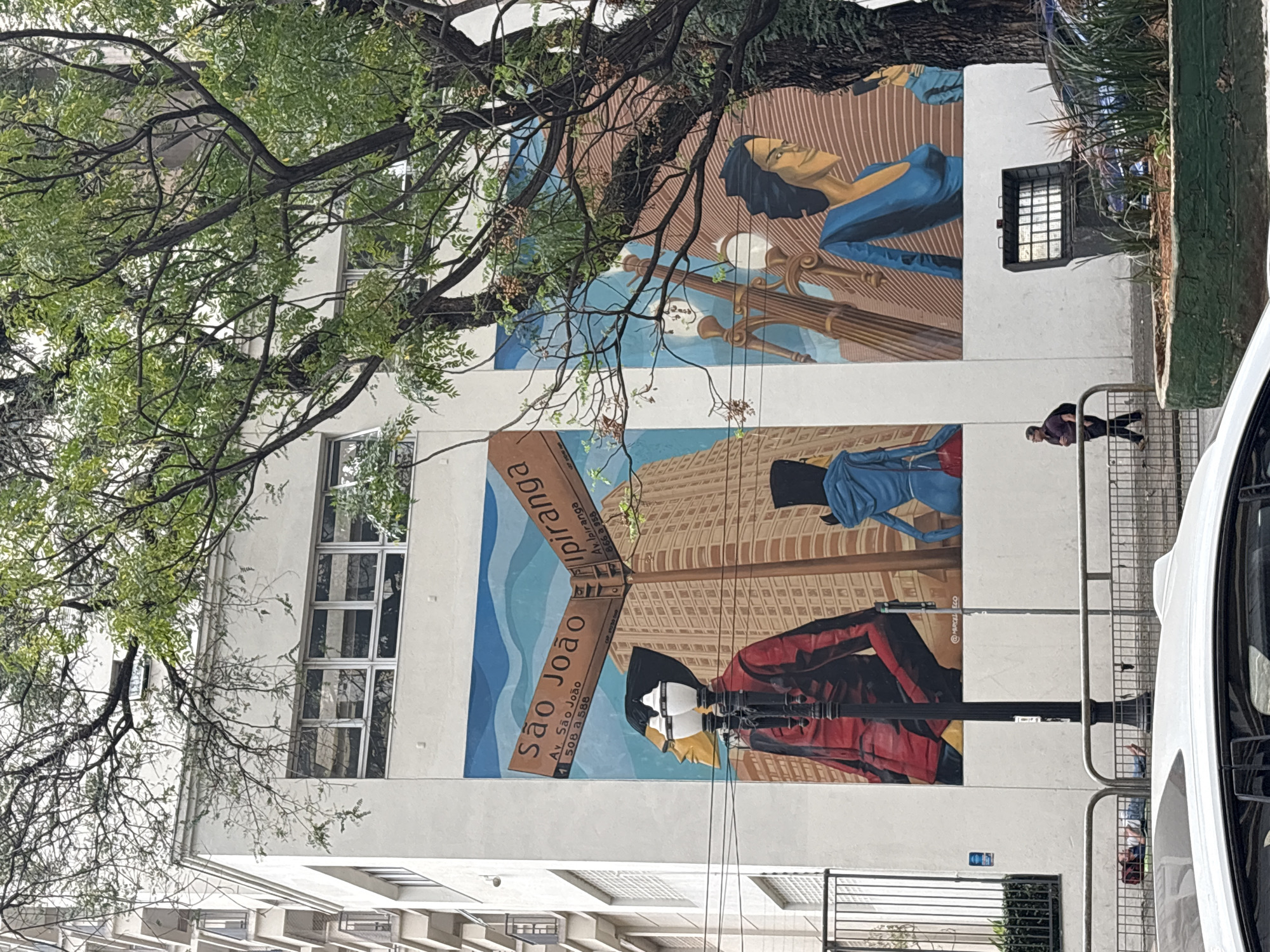 The image depicts a mural on the side of a white building in São Paulo, Brazil. The mural features two street signs at an intersection, one pointing to "São João" and the other to "Ipiranga." Below the signs, stylized figures are painted. On the left, a figure in a red jacket and yellow head is depicted, and to the right, figures in blue clothing are shown. In front of the mural, a lamppost stands, and further to the right, a planter with vegetation and a trash receptacle are visible. A man in a purple shirt and dark pants is walking on the sidewalk to the left of the planter. In the foreground, a white car is partially visible, and a person is lying down on the ground behind a metal fence. Trees with green leaves and branches are in the foreground and partially obscure parts of the building and mural. The building itself has windows with metal frames. The overall scene suggests an urban environment with art integrated into the city's architecture.