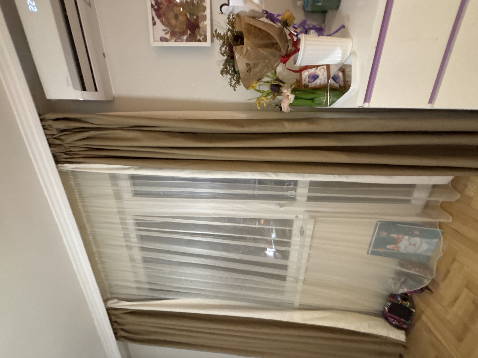 The image shows an interior scene, likely a room, with a window covered by curtains. The curtains are a combination of a heavier, brown-toned fabric at the top and bottom, and a sheer, lighter-colored fabric in the middle. An air conditioning unit is mounted on the wall near the top left corner. There are decorations on a surface above the curtains, including a framed picture and a bouquet of flowers in a vase. A closet is partially visible on the right side of the image with a white front and purple accents. The floor is wooden and appears to be made of planks. The angle and perspective suggest the photo was taken from a standing position inside the room.
Without additional information, such as street names, landmarks, or specific architectural details, it is impossible to determine whether the image was taken in the city of Sofia, Bulgaria.