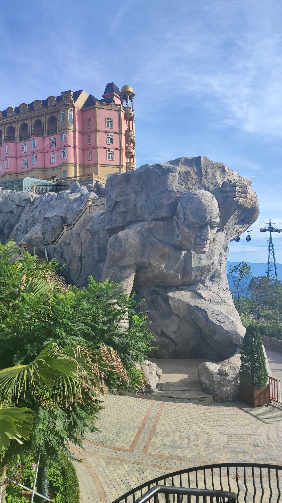 This is a photograph of the Bà Nà Hills complex in Đà Nẵng, Vietnam, specifically showcasing the "Giant's Hand" rock formation with a large carved face and body, which appears to be supporting a rocky overhang. To the left of the rock formation, a large, ornate pink and yellow building with multiple stories, arched windows, and a golden dome is visible, perched on a rocky cliff. Lush green foliage, including palm trees and other plants, frames the foreground and sides of the image. The ground is paved with interlocking bricks in a pattern of tan, grey, and orange. In the background, a cable car station and tower can be seen against a bright blue sky with scattered clouds. The overall scene depicts an architectural and natural landscape designed for tourism.