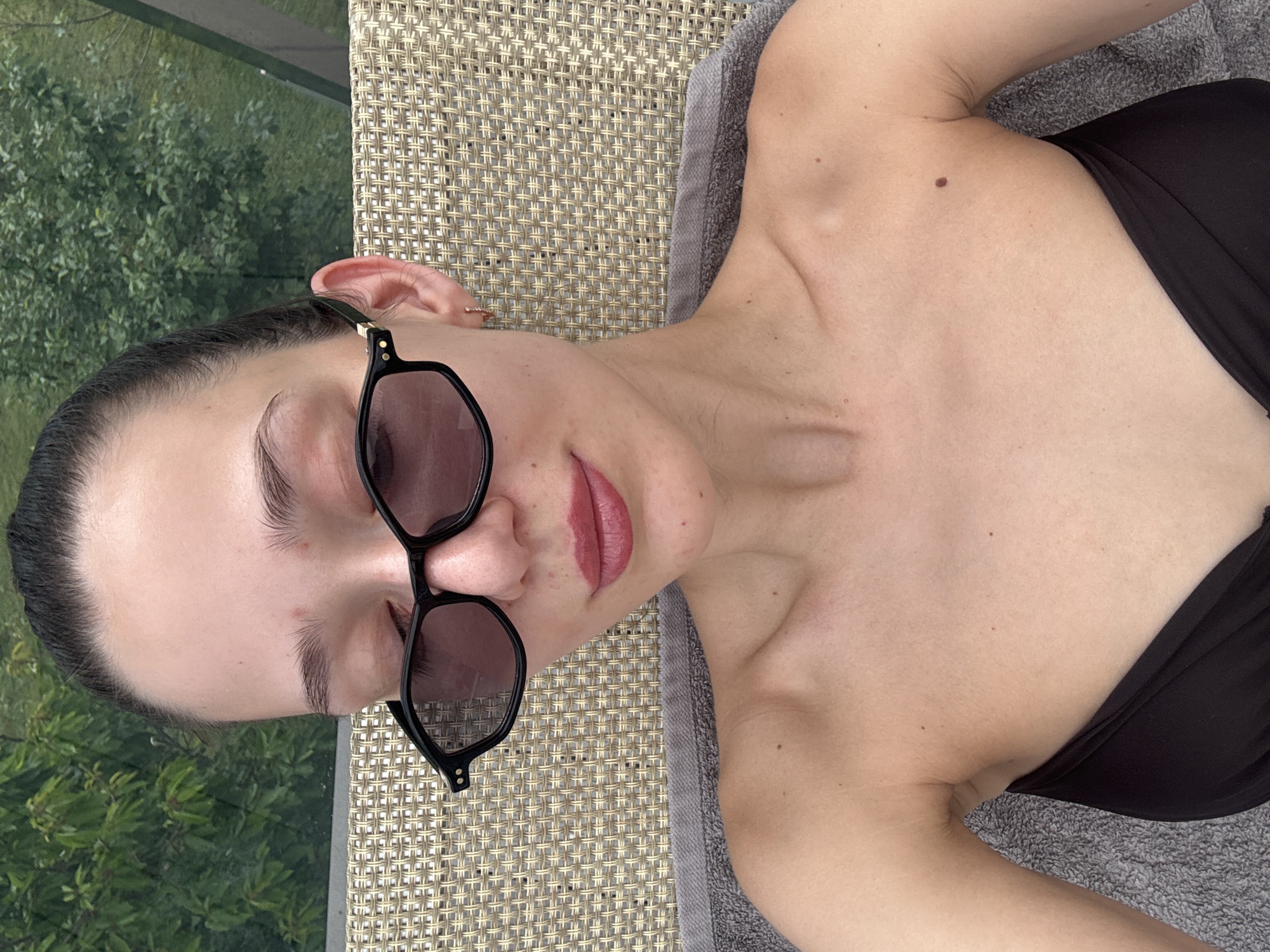 A young woman is lying on a woven outdoor lounge chair in Boreti, Montenegro. She is wearing black sunglasses and a black top. A gray towel is beneath her. Outside the lounger is greenery.
