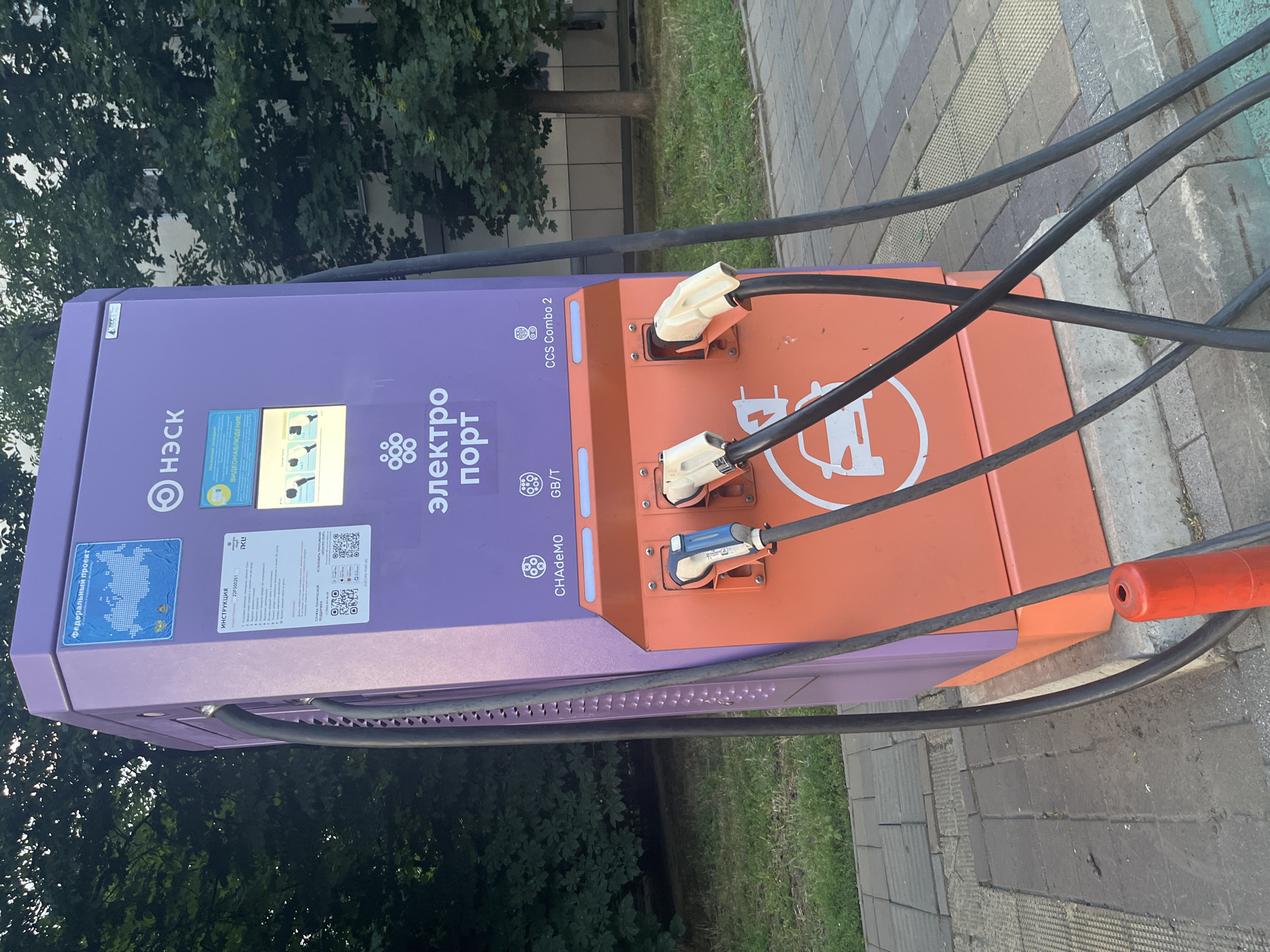 A purple and orange electric vehicle charging station is pictured outdoors on a paved sidewalk next to a grassy area and trees. The charging station has three charging connectors extending from its front, two of which are connected to cars. Text on the charging station includes "Электро порт" and various logos and instructions in Russian. The environment appears to be a city street or public area. No people are visible in the image.