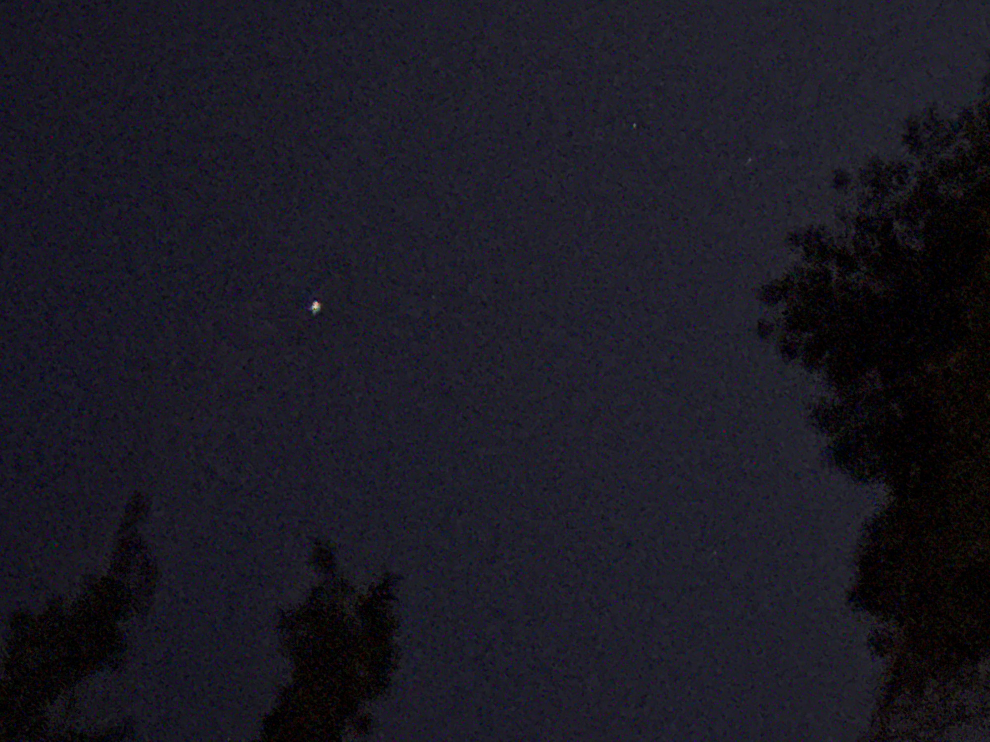 A dark, grainy photograph depicts a night scene with a single bright point of light in the upper left portion of the frame. The remainder of the image is predominantly a dark blue-black color, suggesting a night sky or similarly dimly lit environment. The outlines of trees or other foliage are visible in the lower and right portions of the image, rendered in dark shades and lacking clear detail. The photograph suggests an outdoor setting at night with minimal lighting. It is not possible to identify the location as Sofia, Bulgaria, or any other specific location, without additional contextual information. There is no information regarding people, subjects, objects, actions, or interactions provided within the image itself.