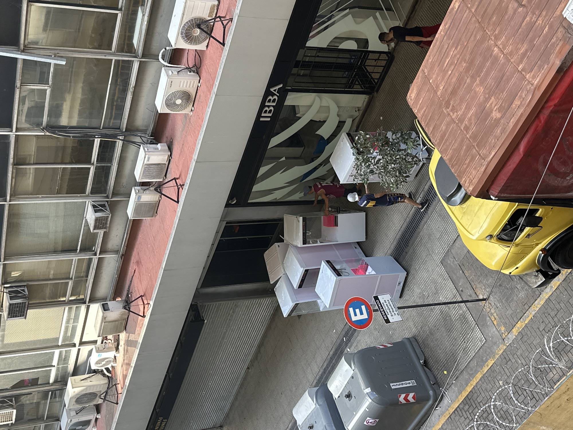 A high-angle photograph depicts a street scene in Buenos Aires, Argentina. Along the left side, an office building with numerous window panels and multiple air conditioning units mounted on its facade. An awning extends from the building with "IBBA" signage. Below the awning, a row of display cases with white frames and glass tops are arranged on the sidewalk. Two men are visible near the cases. Adjacent to the display cases, there is a 'Reserved' parking sign. Further down the street, trash bins are located near the sign. A yellow truck with a red and brown cargo covering parks on the street. The street is paved with concrete squares and edged with razor wire. Two people stand on the sidewalk outside the building.