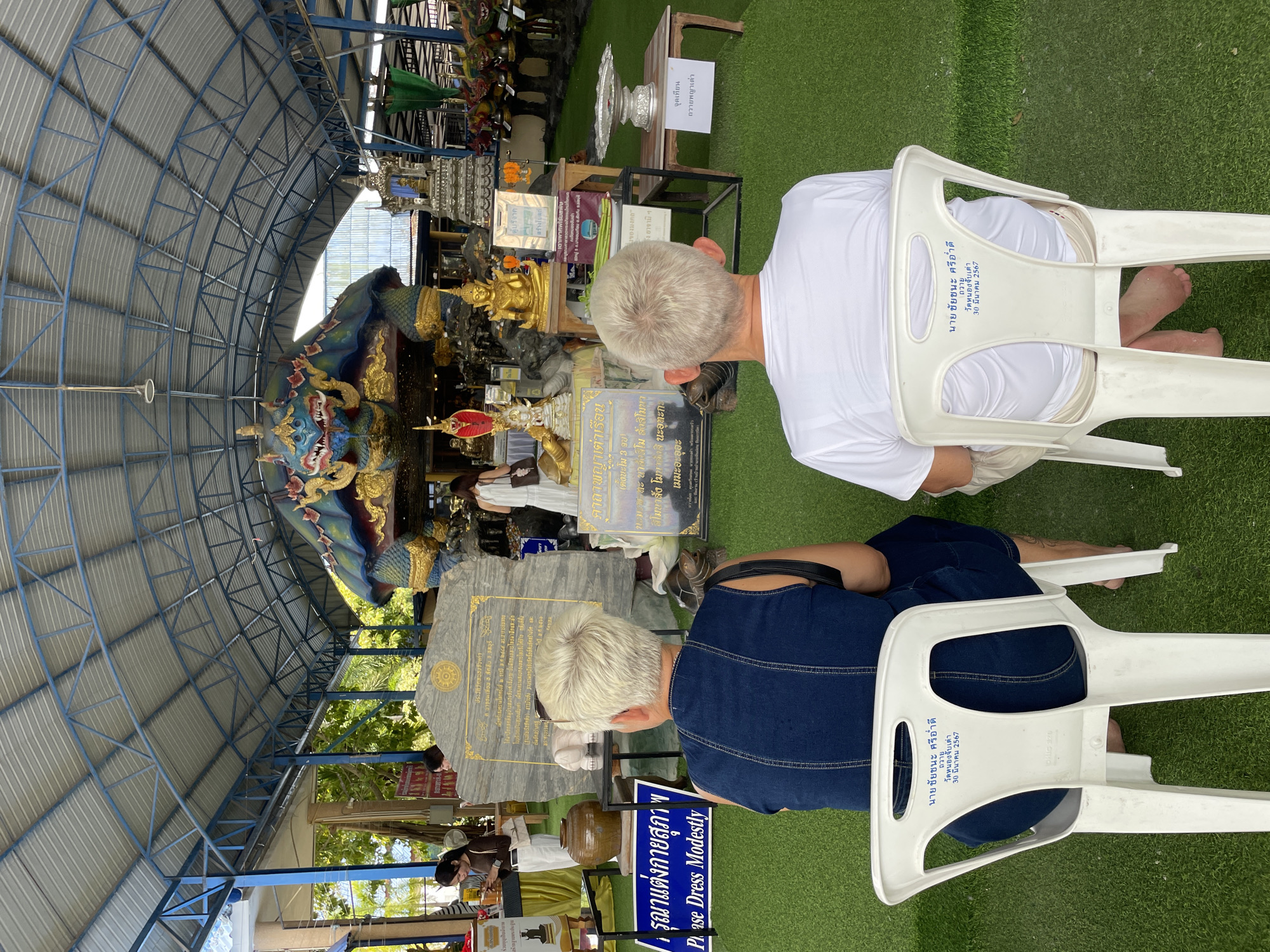 The image shows two individuals, seen from behind, seated on white plastic chairs facing a colorful, dragon-like statue within a covered outdoor structure. The structure has a metal roof supported by blue beams, and the ground is covered with artificial green turf. In the foreground, the back of the chairs has text printed on them, indicating names and a date. The person on the left wears a dark blue denim top, while the person on the right wears a white t-shirt.
Behind the seated individuals, a large stone monument with text in Thai is visible, along with various statues and religious offerings. Further back, other people are present, interacting with the offerings or observing the statues. The overall scene appears to be a religious or cultural site in Bang Sare, Thailand. The presence of Thai script on the monument and the chairs, along with the distinct architectural style and religious imagery, confirms the location. The environment is well-lit, suggesting daytime. The atmosphere seems peaceful and reverent.