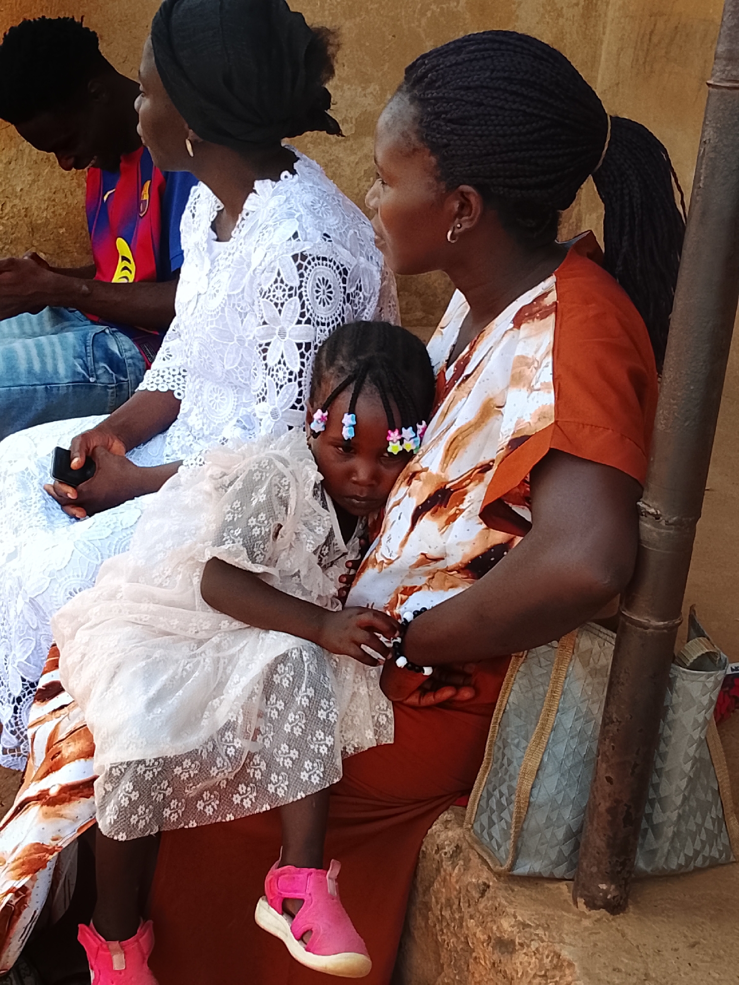 A group of four individuals, likely a family, are seated outdoors in Akwanga, Nigeria. On the far left, a man wears a blue and red striped t-shirt (resembling an FC Barcelona jersey) and blue jeans, looking down. Next to him, a woman in a white lace top and a black head covering holds a smartphone, her face partially visible. On the right, another woman, with braided hair tied in a ponytail and hoop earrings, wears an orange and white patterned top and a rust-colored skirt. She cradles a young girl in her lap. The girl wears a white lace dress, pink sandals, and has braided hair adorned with colorful beads or clips; her expression appears pensive.
The subjects are seated against an earthy-toned wall or structure. A light blue or grey woven tote bag with gold straps rests beside the woman on the right, next to a rusty metal pole. The woman holding the child wears a black and white beaded bracelet on her wrist. The scene appears to be set during the daytime, with soft natural light, suggesting warm weather. The group seems to be relaxing or waiting. No clear readable text is visible, though the man's shirt has a logo.