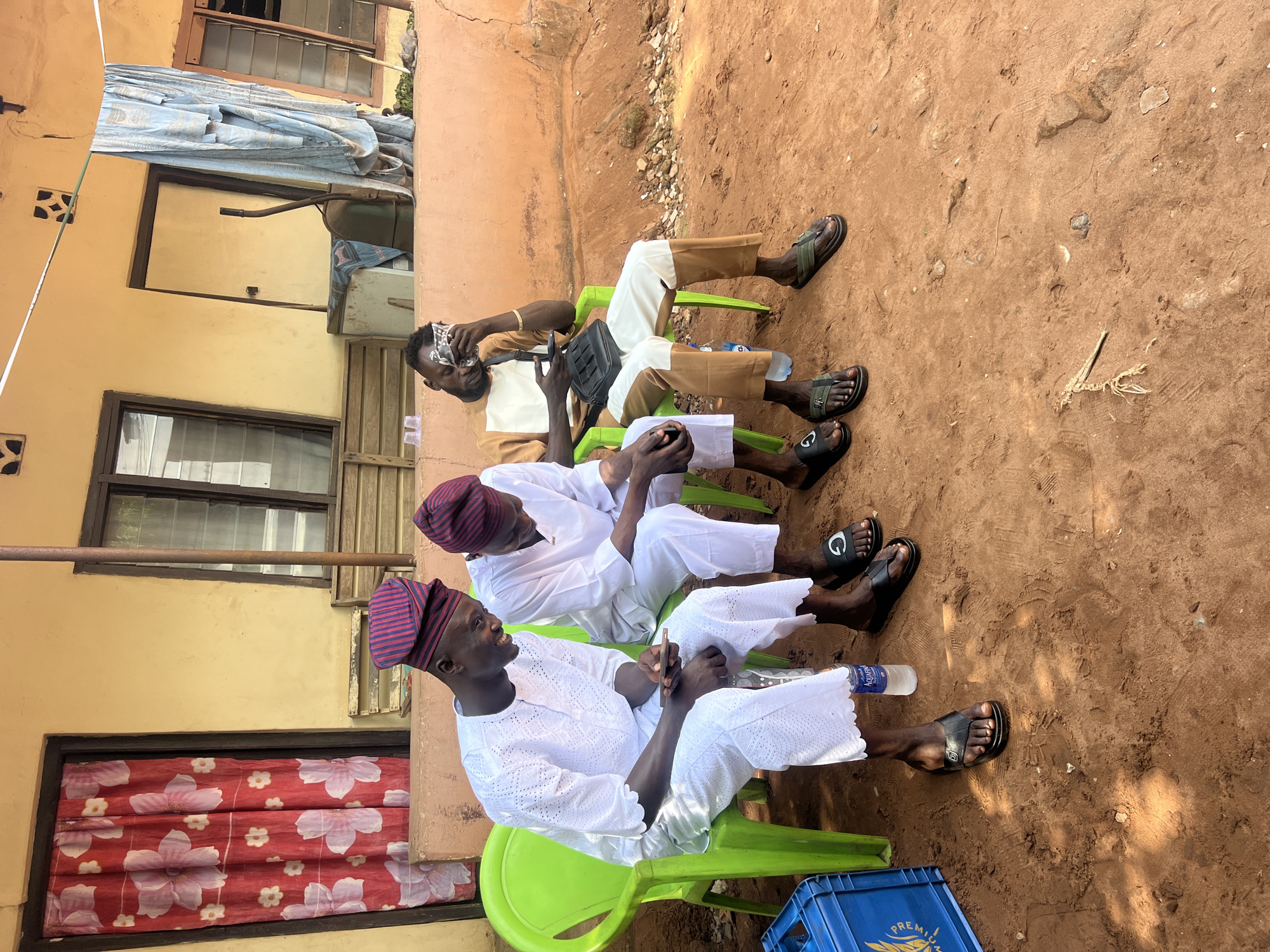 Three men are seated on green plastic chairs in an outdoor setting in Obosi, Nigeria. The first man, nearest the camera, wears a white, lace-textured outfit with a traditional red and purple striped cap. The second man also wears a white outfit and matching cap. The third man wears a tan and white outfit and is holding what appears to be currency in his left hand and a black bag in his right hand. The ground is reddish-brown dirt. Behind the men are the exterior walls of buildings. A blue plastic crate is partially visible at the bottom right corner of the frame.