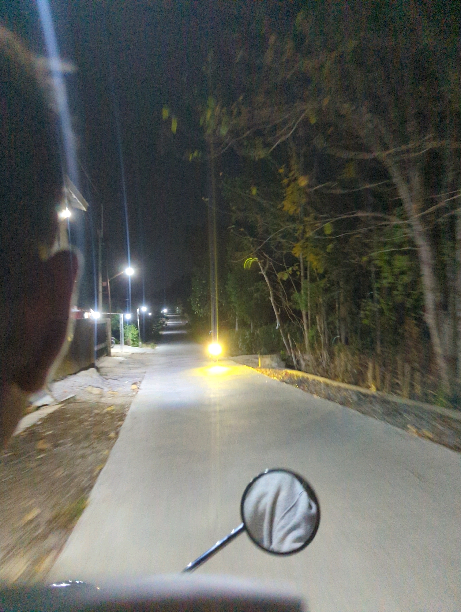 The image depicts a dimly lit, paved road at night, likely in a rural or suburban area of Senggrong, Indonesia, judging by the presence of palm trees and tropical vegetation. The perspective is from someone riding a motorcycle, as indicated by the handlebars and a rearview mirror reflecting a light-colored garment, possibly a hoodie. Streetlights illuminate the road, casting long shadows and creating a sense of depth. The road appears to be a straight stretch, bordered by vegetation on the right and a sidewalk and low structures on the left. The overall atmosphere is calm and nocturnal, with no other vehicles or people visible.