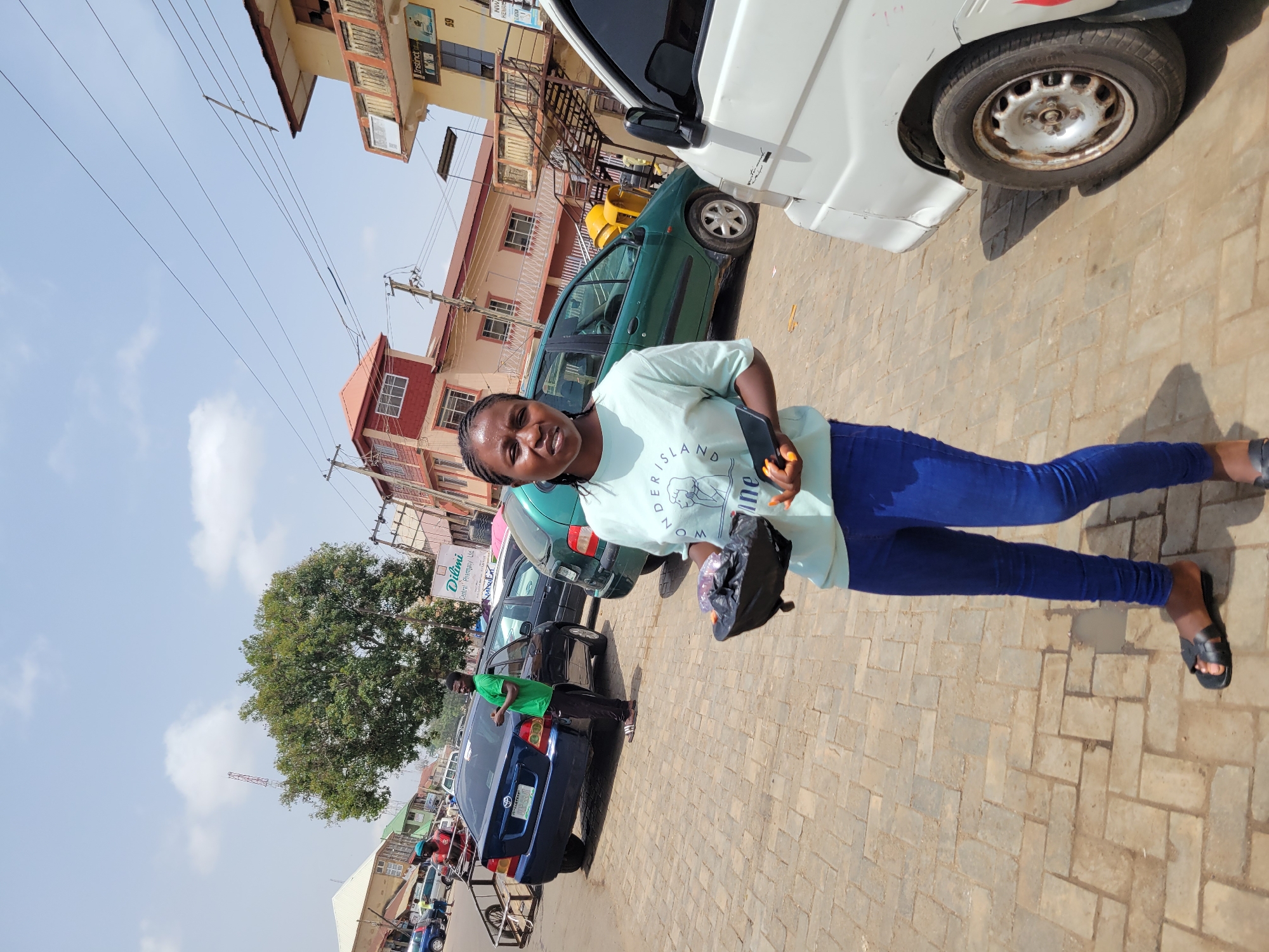 A woman stands on a brick-paved street in Jos, Nigeria. She wears a light-colored T-shirt, blue jeans, and black sandals. She holds a black bag in one hand and a mobile phone in the other.
Behind her are parked cars of different colors, including green, black, and blue. A white van is partially visible on the right side of the image. Buildings are visible in the background. Overhead electrical wires are prominent against the sky. A tree is on the left. A sign of Dilimi Central Pharmacy Ltd is visible in the background. Another person is standing by a car next to the woman. Other people and cars are visible in the distance.