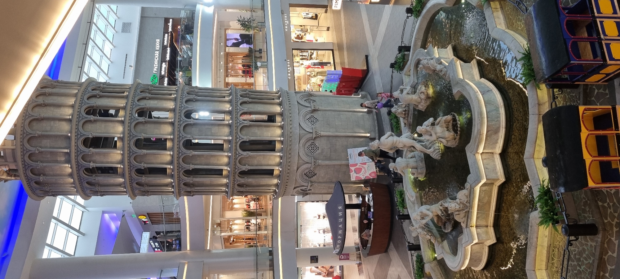 The image captures an indoor scene, likely a shopping mall, with a prominent, decorative replica of the Leaning Tower of Pisa as the central feature. The tower is illuminated, highlighting its architectural details. In front of the tower, a multi-tiered fountain with classical statues is active, with water flowing.
Surrounding the fountain area, there are various elements of a commercial or public space. To the left of the fountain, a circular information booth with the word "ИНФОРМАЦИЯ" (Information) is visible, with two individuals behind the counter and one person approaching it. To the right of the fountain, there are several people, including a woman with a child, and a man interacting with a display. In the background, multiple levels of storefronts are visible, with some displaying merchandise and illuminated signage. One sign reads "ГРУЗИНСКАЯ КУХНЯ" (Georgian Cuisine). The upper sections of the mall show a ceiling with large windows or skylights and artificial lighting, including blue accent lighting. At the very bottom of the image, two colorful, toy train carriages are positioned. The overall environment suggests a well-lit, busy indoor public space designed for shopping and leisure, potentially part of a larger entertainment complex. The scene is set in Moscow, Russia, at the Vegas Crocus City mall.