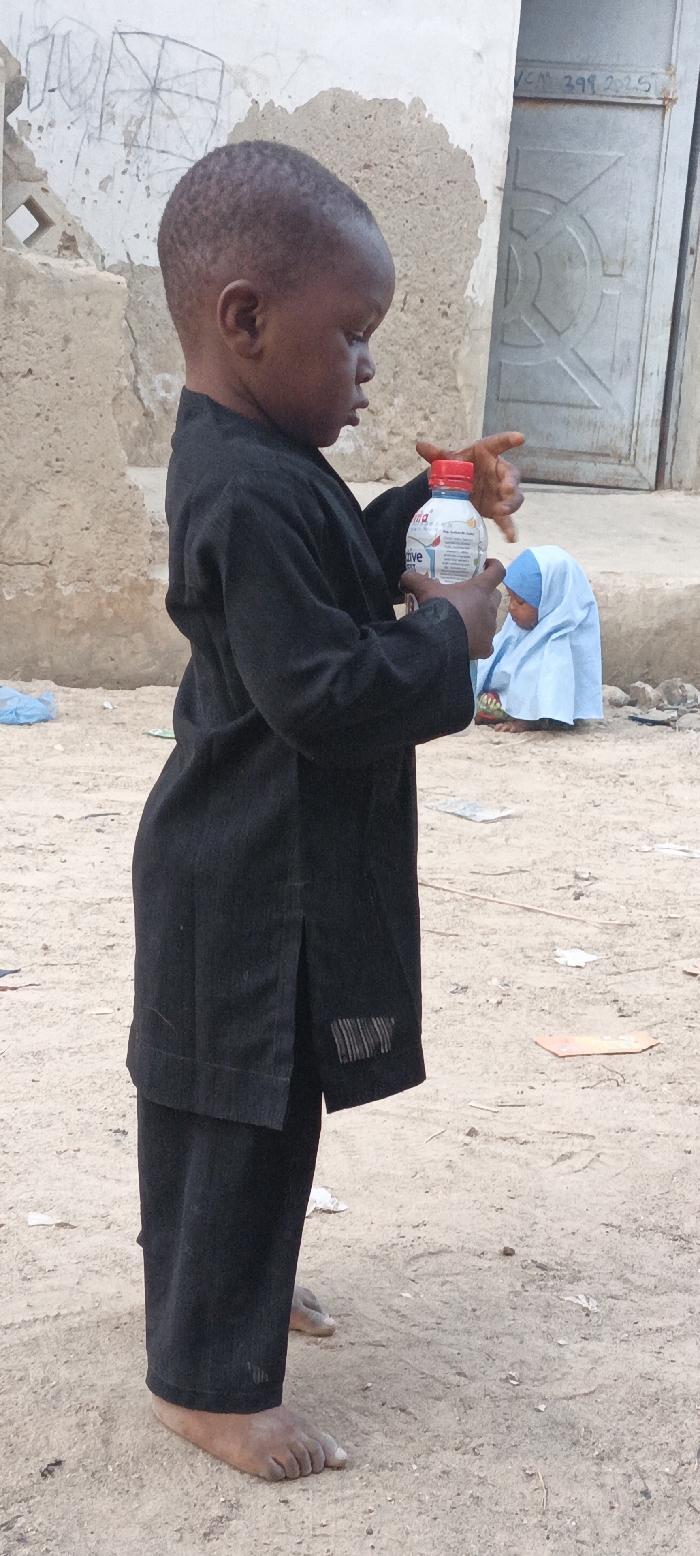 A young child, likely male, dressed in a black long-sleeved tunic and trousers, stands barefoot on sandy, light-brown ground in Rangaza, Nigeria. The child is captured in profile, facing towards the right side of the frame, intently looking down at a plastic water bottle held in both hands. The bottle has a red screw cap and a label prominently featuring the word "active" along with a colorful bird graphic.
In the mid-ground, a smaller child, partially obscured, is seated on the ground, wearing a light blue head covering. The background consists of weathered, light-colored walls; one wall behind the main child's head shows a faint, rough drawing resembling a square with diagonal lines. Further back and to the right, a grey metal door with an embossed circular pattern is visible, bearing faded text that appears to read "VCN 399 2025" near the top right corner. The ground is uneven dirt or sand, scattered with small pieces of debris. The scene is illuminated by natural daylight, indicating it is daytime. The overall setting suggests a humble, possibly rural or semi-urban environment.