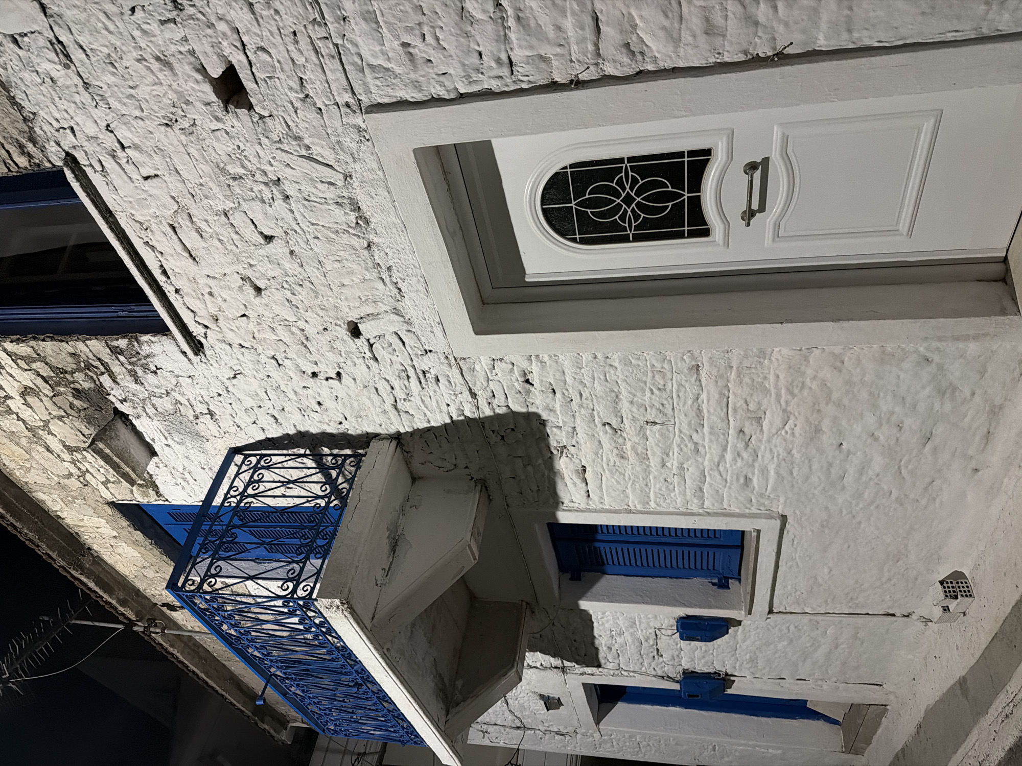 The image depicts a nighttime exterior view of a white brick building with blue shutters and a blue wrought-iron balcony railing. The building is located in the Municipal Unit of Alyzia, Greece. A white front door with a decorative glass pane is visible on the right side of the image. To the left, a blue door is slightly ajar, and a blue mailbox is mounted on the wall. Above the blue door, a window with blue shutters is present. The balcony, painted blue, extends from an upper floor and is adorned with intricate metalwork. The rough texture of the white brick walls is prominent. The scene is illuminated by artificial light, casting shadows on the building and the street. A small pile of red bricks is on the ground near the lower left corner. An antenna is affixed to the rooftop.