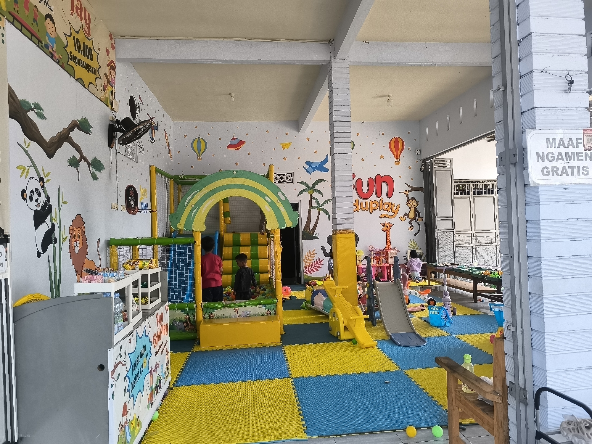 The image depicts a brightly decorated indoor playground, likely a children's play center, located in Dagangan, Indonesia. The walls are adorned with colorful murals featuring cartoon animals, hot air balloons, and various playful motifs. The floor is covered with interlocking foam mats in blue and yellow.
In the center of the scene, a multi-level play structure with a green and yellow canopy and a slide is visible. Several children are present. Two boys, one wearing a red shirt and the other a black shirt, are interacting with the play structure. Another child, wearing a purple hoodie, is seated at a table in the background. In the foreground, a grey counter is visible, with shelves containing snacks and drinks. A white sign with black text is mounted on a white paneled wall on the right side of the image, reading "MAAF NGAMEN GRATIS."
The overall environment suggests a safe and engaging space for children to play and socialize. The play equipment is designed for active recreation, with slides, tunnels, and a ball pit. The colorful decorations and cheerful atmosphere contribute to the inviting nature of the establishment. The presence of security cameras indicates a concern for safety and monitoring within the facility.