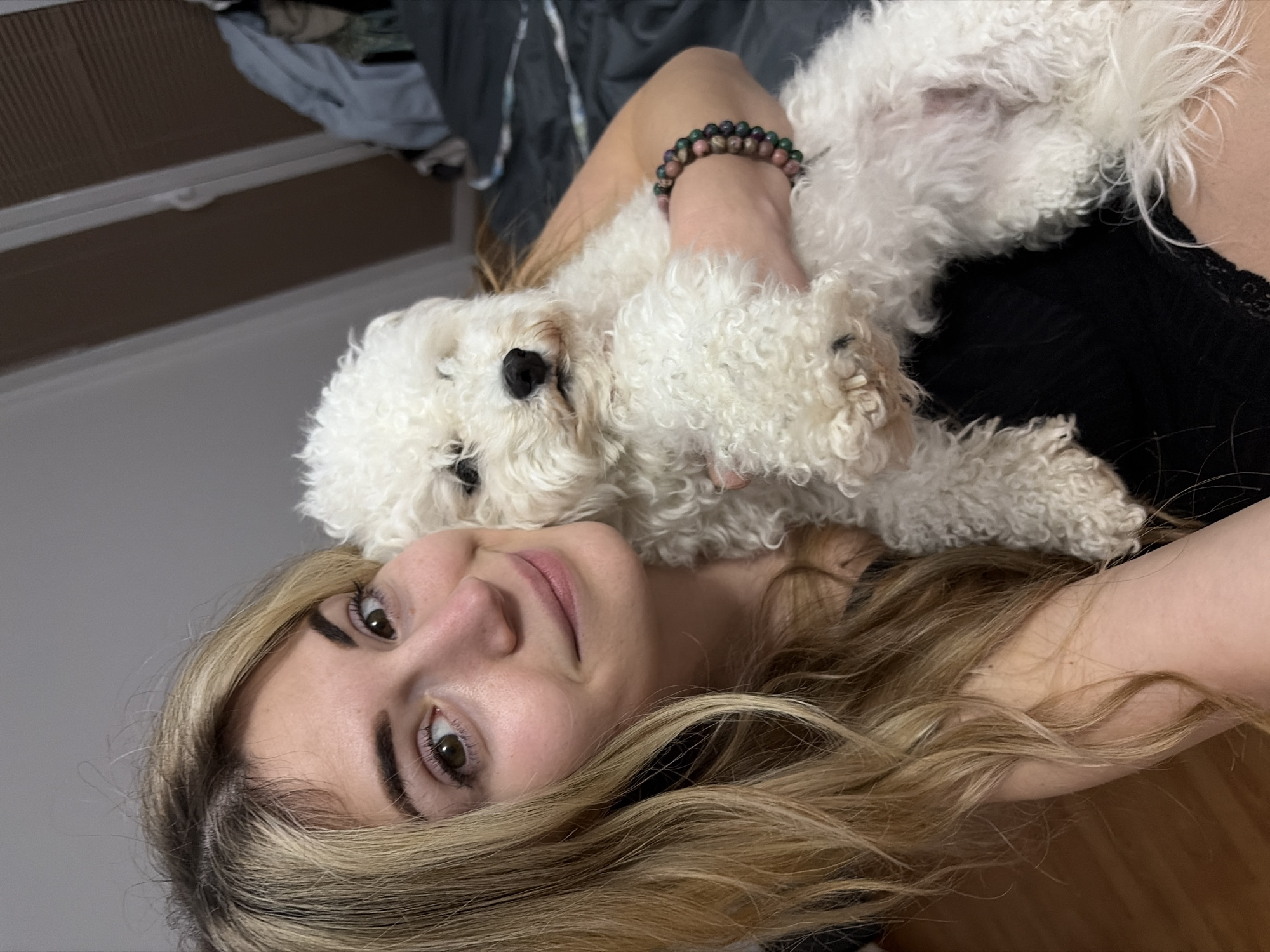 The image displays an indoor scene featuring a young adult female and a small white dog. The female, characterized by long blonde hair and brown eyes, is positioned centrally, looking towards the camera with a subtle smile. She wears a dark garment, potentially black, and has a multi-colored beaded bracelet on her right wrist, composed of dark green and reddish-pink beads. She is holding a white, curly-haired dog, possibly a Bichon Frise, with dark eyes and nose, which is also oriented towards the camera. The immediate background shows a light-colored wall. To the right, a dark grey bedding or fabric item is visible, along with a light blue textile object. An angled window or internal partition, fitted with a brown blind, is partially visible in the upper right quadrant. The lower left foreground reveals light-toned wooden flooring. The environment suggests a residential or hotel room setting. No specific landmarks or unique architectural features are present in the image to identify a precise location within Budva, Montenegro.