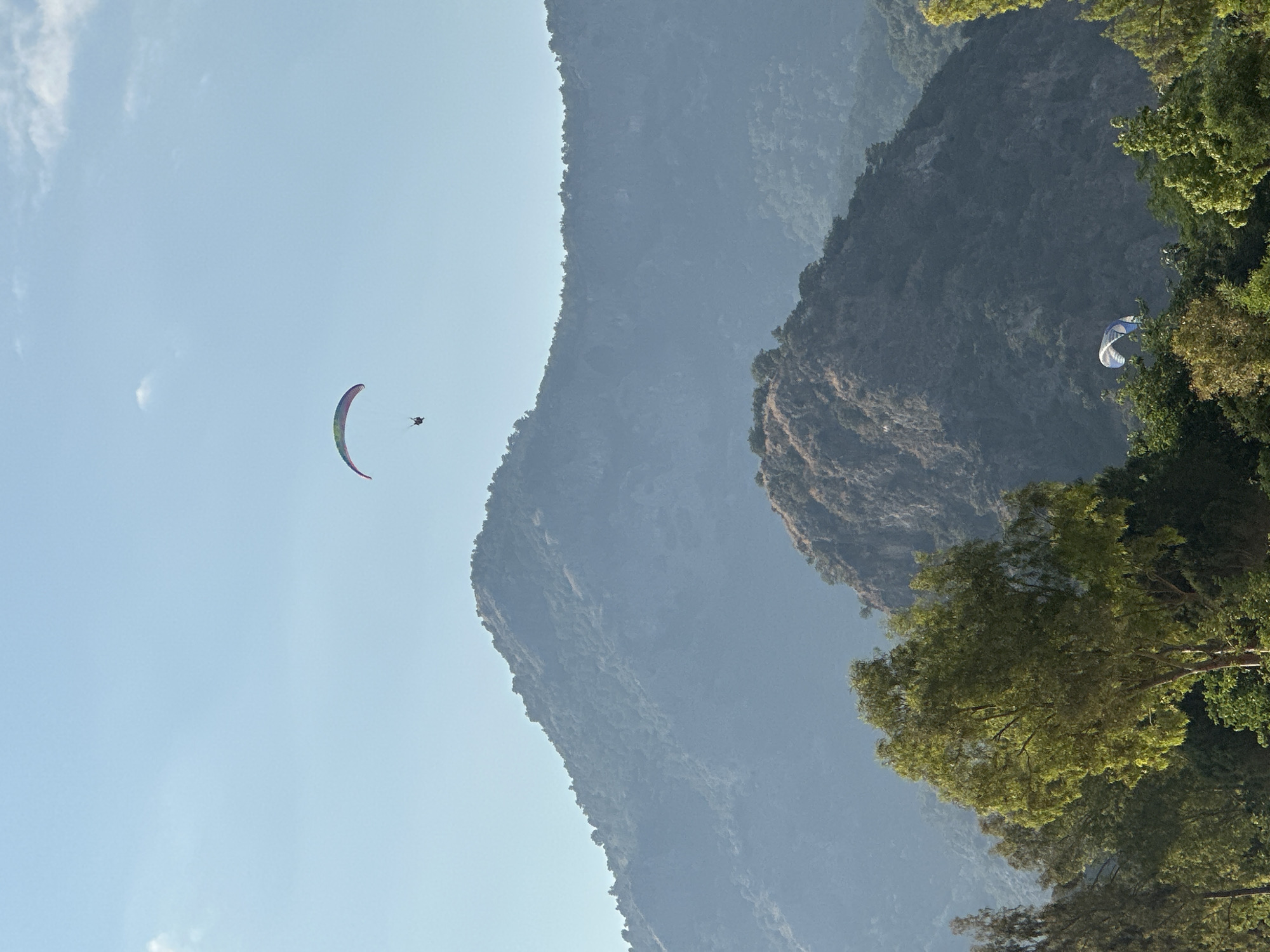 This daytime photographic image depicts a scenic landscape in Fethiye, Turkey, featuring mountains, lush vegetation, and two paragliders under a clear sky.
The upper two-thirds of the frame are dominated by a pale blue sky with a few scattered, wispy white clouds, primarily in the upper left quadrant. Centered in the upper portion of the sky is a single paraglider with a vibrant, arc-shaped canopy displaying a gradient of colors including green, blue, purple, and reddish-pink. A single individual in a harness is suspended beneath the canopy.
Below the horizon, a series of mountains recede into the distance, exhibiting atmospheric perspective. The nearest large mountain, occupying the midground, is covered in dense, dark green foliage and appears hazy with a bluish-grey tint. Its peak is visible on the left, with its ridge extending towards the right. Further behind, additional mountain layers are visible, becoming progressively lighter and bluer, almost blending with the sky.
In the lower third of the image, the immediate foreground is characterized by dense, verdant foliage. Prominent green trees, some illuminated by direct sunlight on their leaves, are visible on the left and right sides. Behind these foreground trees and to the lower right, a second paraglider with a blue and white striped canopy is partially visible, appearing at a lower altitude than the primary paraglider and partially obscured by the trees and mountain contours. The overall scene is brightly lit by natural sunlight, suggesting clear weather conditions.