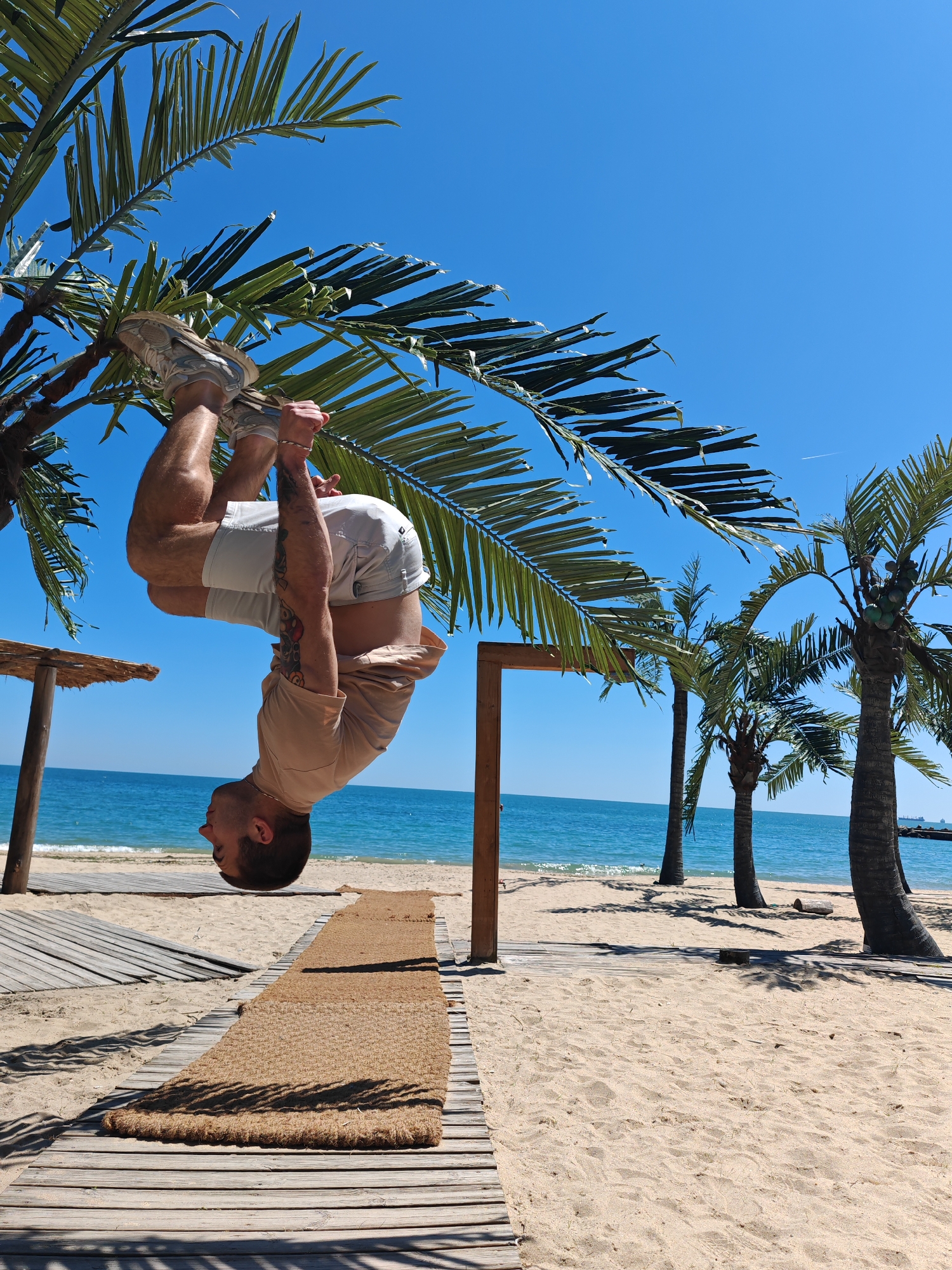 A man is hanging upside down from a palm tree on a beach in Varna, Bulgaria. He is wearing white shorts and a tan shirt, with sneakers on his feet. He is holding onto the palm tree branches with his hands. The background shows a beach with palm trees, blue water, and a clear blue sky. A wooden boardwalk with a coconut fiber mat is visible under him. Other palm trees dot the shoreline.