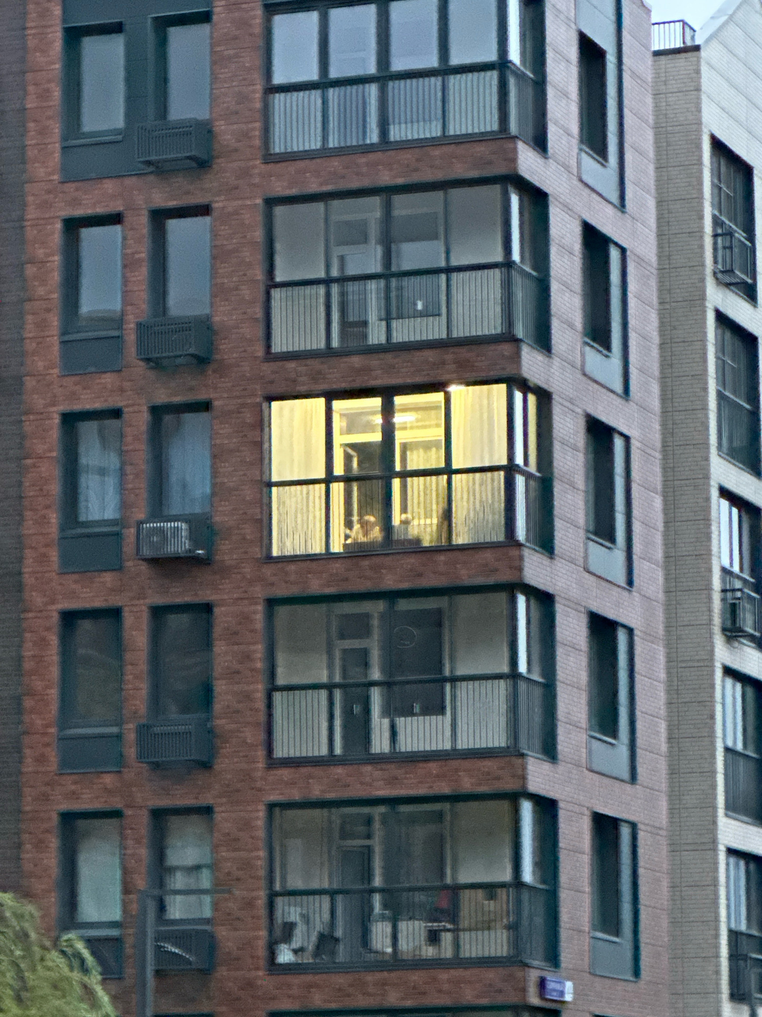 The image depicts a close-up, angled view of the exterior of a modern apartment building, likely in Vidnoye, Russia. The building features a brick facade in a reddish-brown hue, with contrasting dark gray window frames and balconies. Several floors are visible, each with multiple windows and at least one balcony.
On the fourth floor from the bottom, one large window is illuminated from within, revealing a curtained interior with what appear to be two figures sitting or standing. The light spills out, suggesting occupancy. Other windows on this floor and adjacent floors are either dark or show the reflection of the sky.
Adjacent to the brick building is another building with a lighter, possibly gray or beige, brick facade. This building also has multiple floors with windows and balconies. Some windows on this adjacent building are also visible, some dark and some reflecting the sky.
Below the illuminated window on the fourth floor of the brick building, a balcony with a dark railing is visible. Below that, another floor with a balcony and windows is present. On the floors below, more windows and balconies are visible. Some windows have what appear to be air conditioning units mounted beneath them.
The overall impression is a residential urban environment, with the focus on the architectural details and the illuminated interior suggesting a domestic scene within the broader cityscape. The lighting suggests it may be dusk or evening.