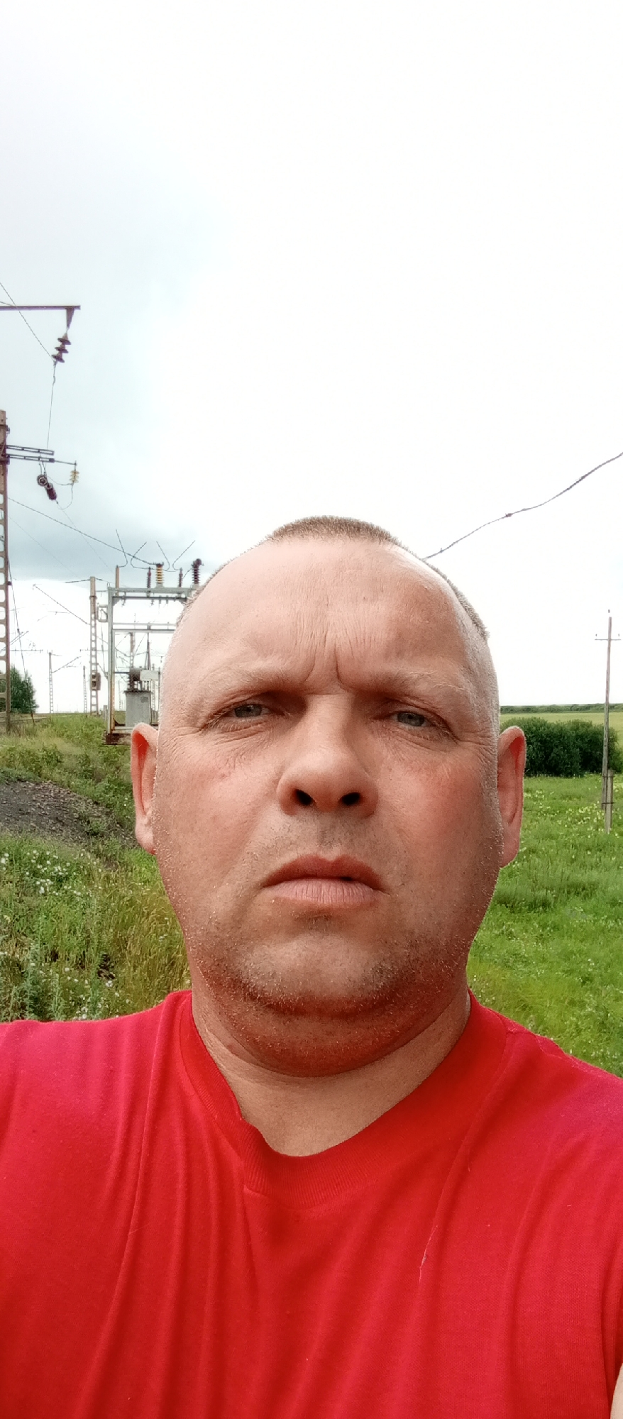 This is a close-up, front-facing selfie of a bald, middle-aged man wearing a red t-shirt. He has a serious expression with furrowed brows. The background suggests an outdoor, possibly rural or industrial setting. To the left, there are power lines and electrical equipment, including what appears to be a transformer substation, mounted on metal poles. This indicates proximity to an industrial or utility area. The ground in front of the equipment is covered with dark gravel or ballast, typical of a railway or industrial site. Beyond this, there is a field of green grass with scattered white wildflowers. In the distance, more green bushes and trees are visible, along with a cloudy, overcast sky. The presence of power infrastructure and the natural landscape suggest a location like Marinovka, Kazakhstan, where such environments can coexist. The lighting indicates it is daytime, but the overcast sky suggests it might be a cloudy day, possibly before or after rain. The man's direct gaze and slightly furrowed brow convey a serious or perhaps pensive mood. No text is visible in the image.