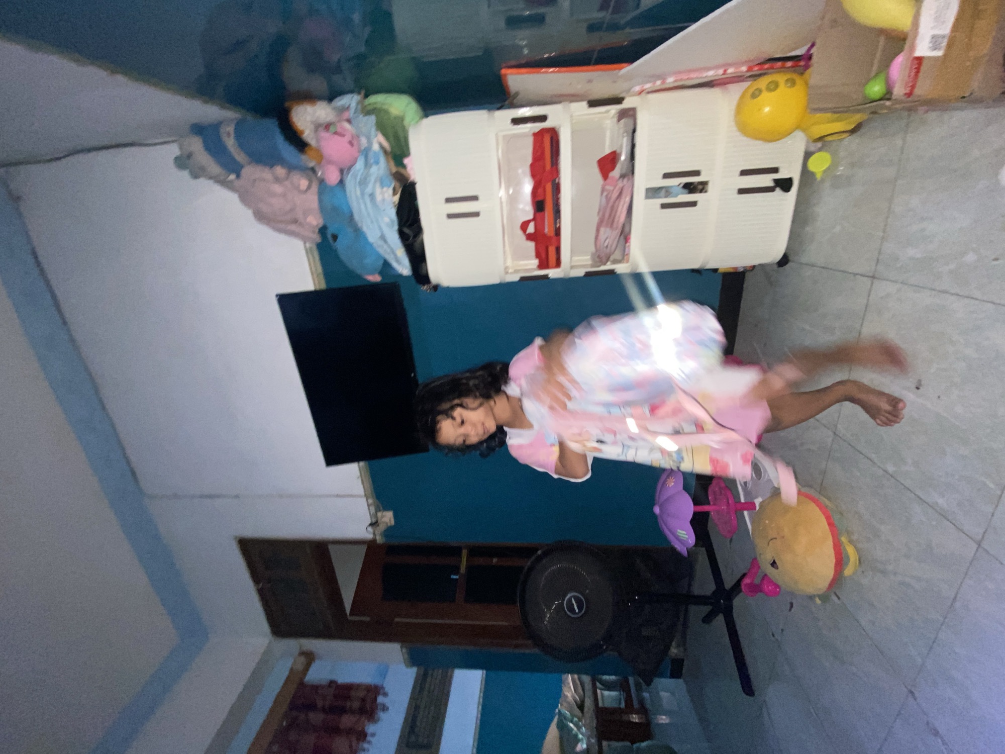In Gelam, Indonesia, an indoor scene depicts a young girl in a pink and white dress, holding a pink and white fabric, likely a blanket or towel, in her right hand. She is standing on a tiled floor, with her left leg forward and bent. To her left, a black oscillating fan stands on a tripod base. In front of the fan and to the girl's left, a large, round, yellow plush toy resembling a burger lies on the floor. A small, purple umbrella toy is positioned between the fan and the burger toy.
Behind the girl and to her right, a tall, white plastic cabinet with three drawers stands. The top drawer contains two red shoes. The middle drawer holds folded pink items. Above the cabinet, various stuffed animals and blankets are piled up. A black flat-screen television is mounted on a blue wall behind the stuffed animals.
Further to the right of the cabinet, a cardboard box contains green and yellow plastic toys, including a yellow duck toy. The room has blue-painted walls with a white ceiling accented by a blue trim. A wooden door with glass panes is visible in the background to the left. The lighting suggests it is either evening or nighttime.