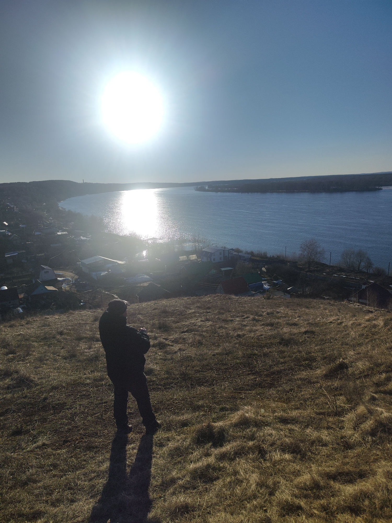 A medium shot depicts a person standing on a grassy slope overlooking a river and a cityscape in Noviy, Russia. The person, clad in dark clothing, faces away from the camera with their arms crossed. The scene is bathed in bright sunlight, which reflects off the water and casts a long shadow of the person onto the dry, brown grass. A cluster of houses is nestled along the riverbank on the left, with an island or distant shoreline visible across the water on the right. The sky is clear and blue.