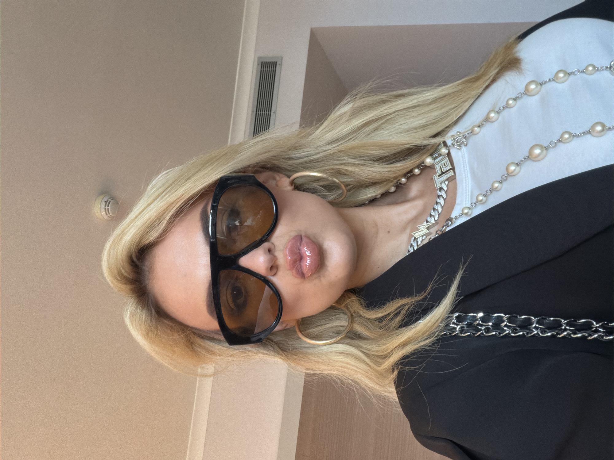 A blonde woman takes a close-up selfie indoors, located in Barcelona, Spain. She is wearing large black-framed sunglasses with amber-tinted lenses, which partially obscure her eyes. Her lips are pursed in a "kiss face" expression and are coated with a glossy, light pink product. She wears large gold hoop earrings. Her attire consists of a black blazer over a white t-shirt. Adorning her neck are multiple necklaces: a chunky silver-toned chain featuring a "CHANEL" pendant with stylized letters, and a long pearl necklace interspersed with silver-toned chain links, which includes a small "CC" logo charm. The silver chain strap of a handbag is visible over her left shoulder. The background reveals a light-colored ceiling with a white circular smoke detector and a rectangular air vent. A subtle white trim runs along the top of the wall on the left side of the frame.