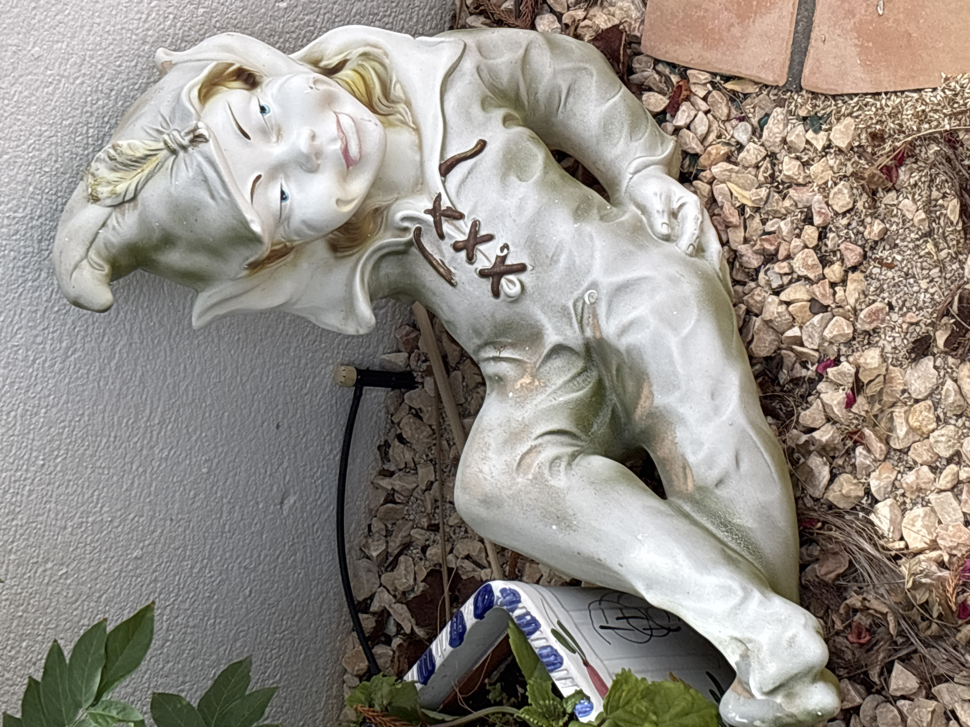 The image shows a garden statue of a gnome lying on its back. The gnome is wearing a pointed hat with a feather, a tunic with lace-up details, and pants. It has a friendly expression with blue eyes and a slight smile. The statue is resting on a bed of gravel and small rocks, with some dried leaves and a terracotta tile visible. To the left of the gnome, a portion of a potted plant in a decorative ceramic planter is visible. The background is a textured, light-colored wall. The scene appears to be outdoors, likely in a garden setting.