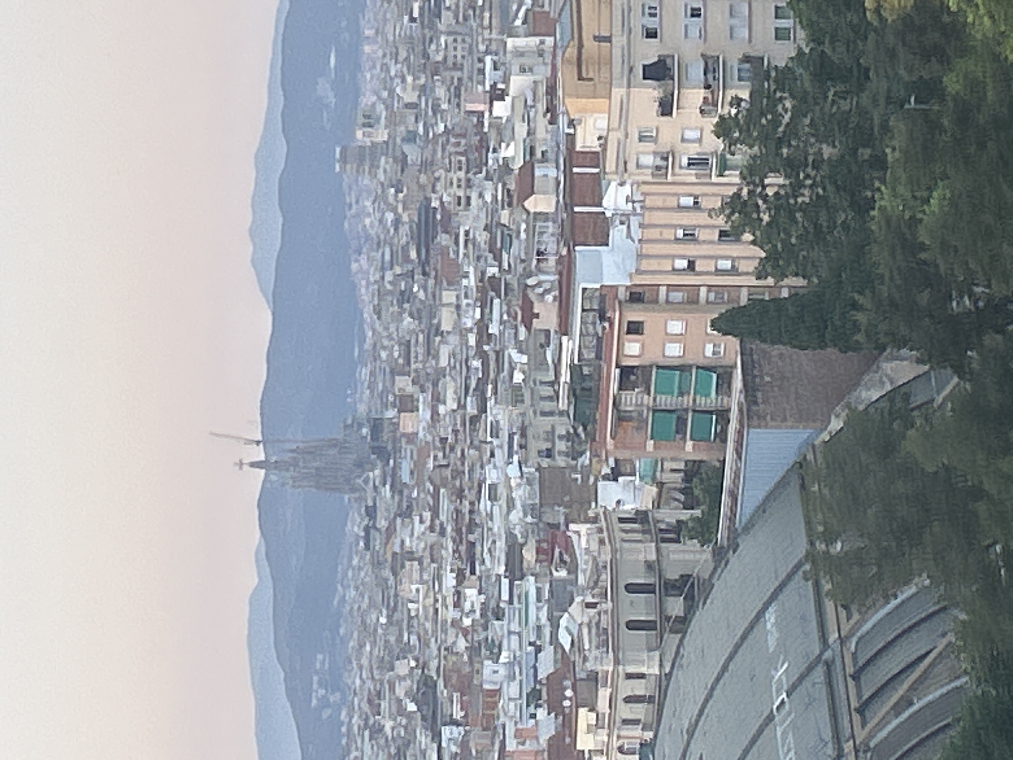 This is a telephoto, eye-level, landscape orientation photograph of a dense urban cityscape under a soft, hazy sky, likely at dawn or dusk. The view is panoramic, showcasing numerous buildings of varying heights and architectural styles, predominantly residential apartments with balconies. The color palette is muted, with soft blues, grays, and light browns dominating the scene, creating a serene and slightly melancholic atmosphere.
In the middle ground, a prominent, iconic church spire with intricate gothic architecture rises above the surrounding buildings. This is recognizable as the Sagrada Familia, Gaudí's unfinished masterpiece, in Barcelona, Spain. Cranes are visible near the spire, indicating ongoing construction.
In the foreground, a large, curved, metallic structure with a ribbed surface and skylights is partially visible on the left. This appears to be a large roof or dome, possibly of a stadium or convention center. Trees, some tall and slender, others bushy, are scattered throughout the foreground and middle ground, adding touches of green to the otherwise urban landscape.
In the background, a series of layered, undulating mountains fade into the hazy sky, suggesting the city's proximity to natural topography. The overall impression is one of a sprawling metropolis nestled between mountains and the sea, with a unique architectural landmark as its focal point. There are no visible people or vehicles in the frame.
